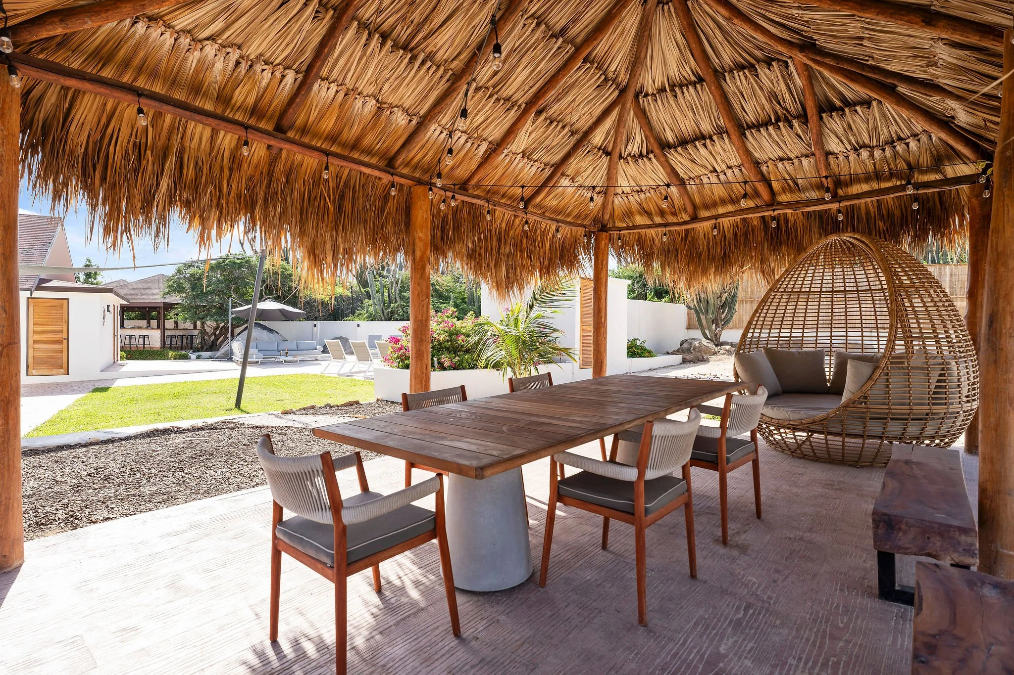 Outdoor gazebo with a thatched roof, wooden tiles, and a large wooden dining table surrounded by chairs. There is a hanging wicker chair on the right, string lights overhead, and a backyard with grass, bushes, and neighboring houses in the background.
