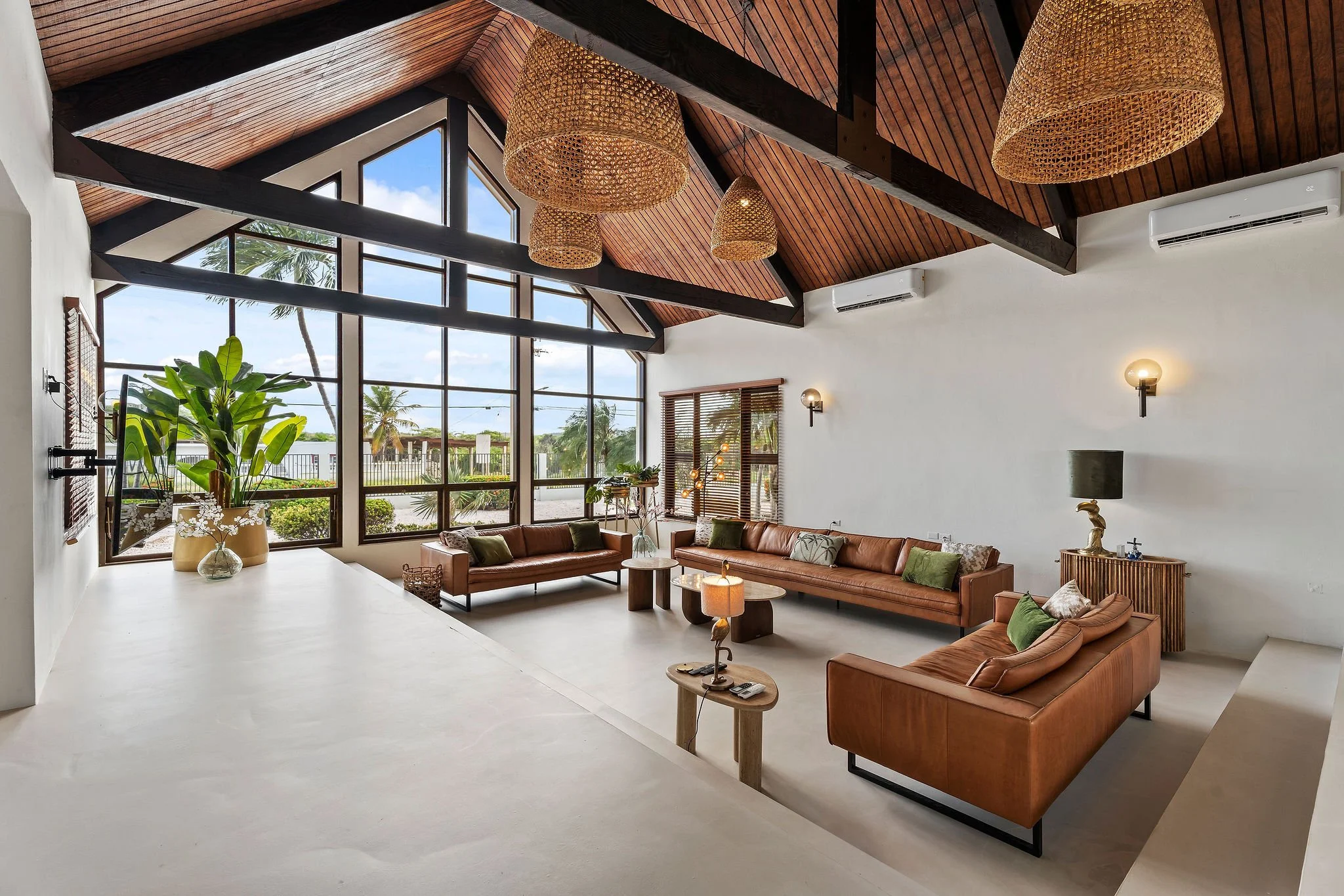 A spacious living room with large floor-to-ceiling windows, wooden vaulted ceiling with black beams, and rattan pendant lights. The room features leather sofas, a TV, a side table with a lamp, plants, and decorative items.