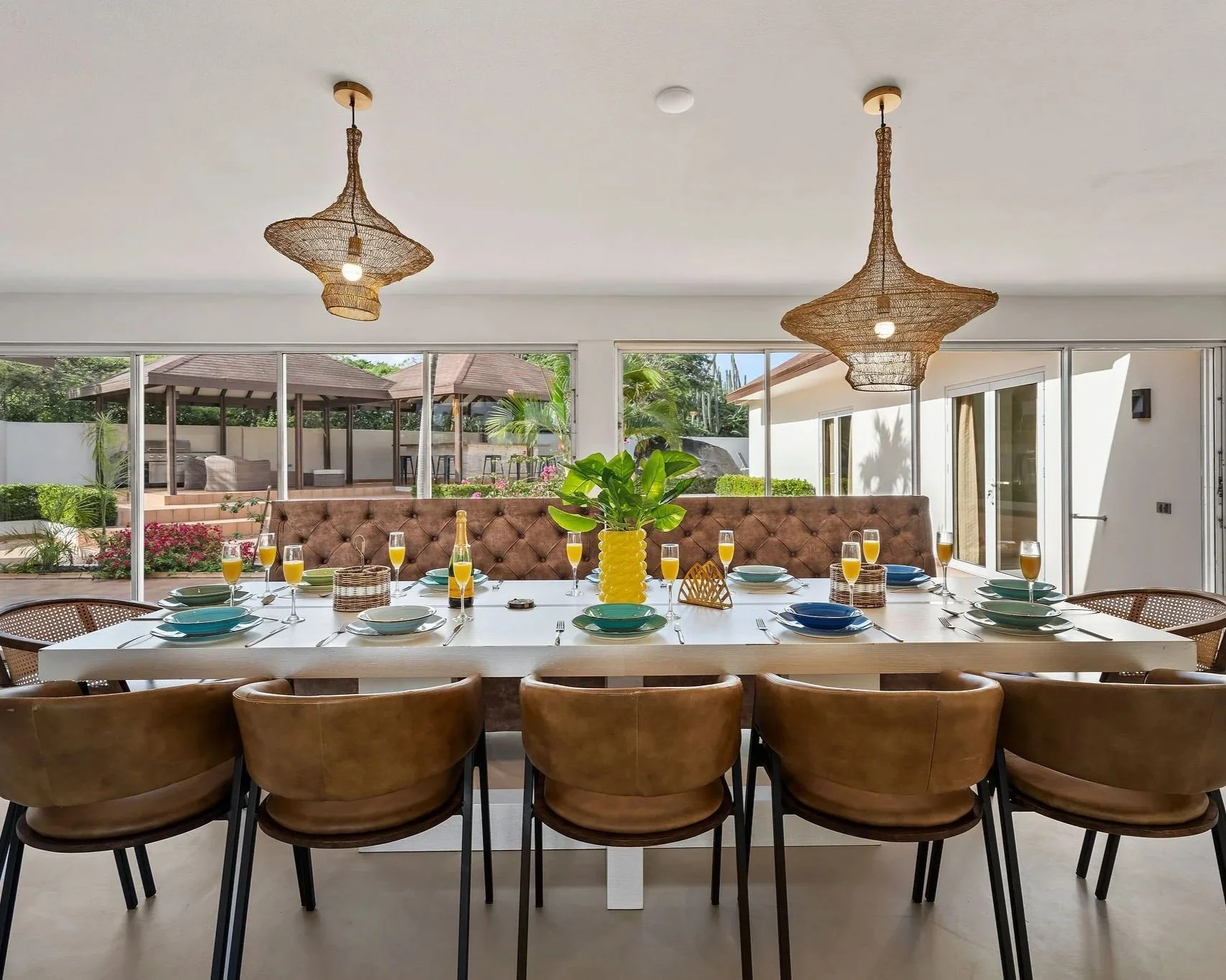 A modern dining room with a large table set for a meal, topped with yellow drinks, green and blue dishes, and a large yellow vase with a leafy green plant. The room has two rattan pendant lights hanging from the ceiling and large sliding glass doors that open to a backyard with outdoor furniture, greenery, and a covered patio.