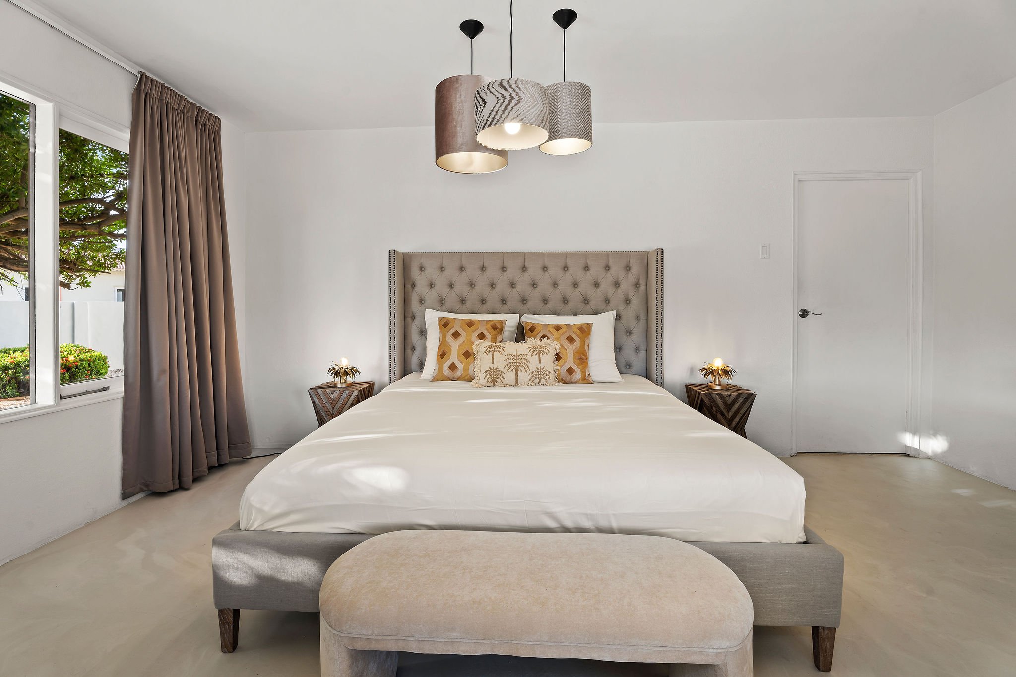 A bedroom with a large bed, beige tufted headboard, and decorative pillows. There is a beige upholstered bench at the foot of the bed, side tables with lamps, a window with beige curtains, and a modern light fixture hanging from the ceiling.