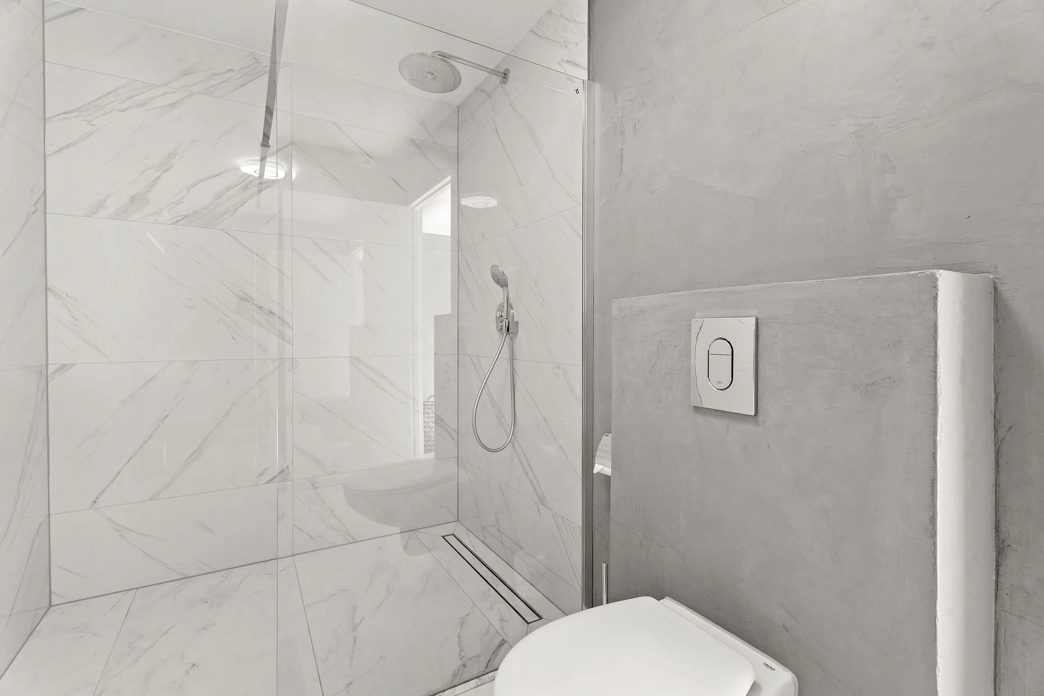 Modern bathroom with white marble tiled shower area, rainfall showerhead, handheld shower, and toilet with concealed cistern.