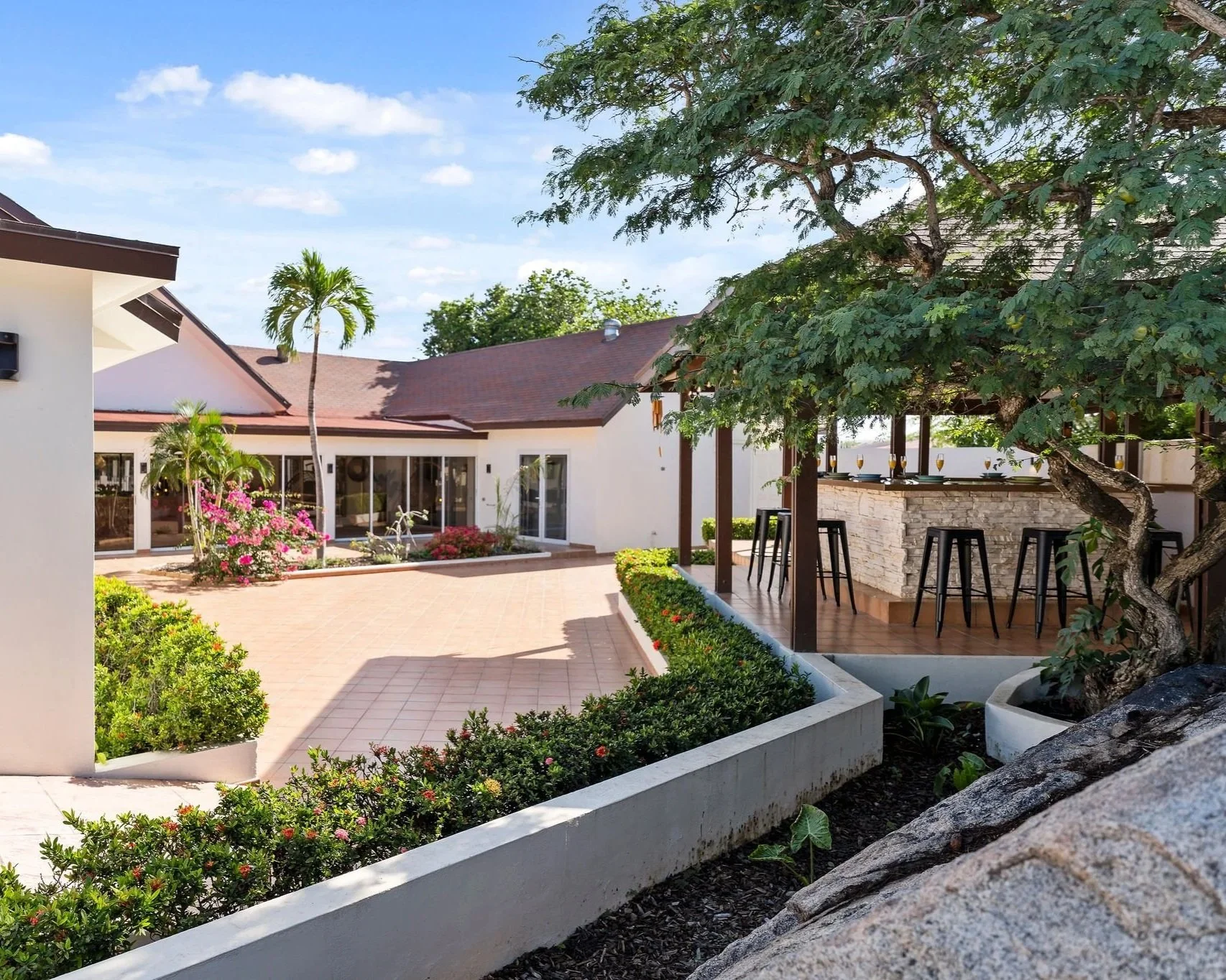 Sunny backyard with a white house, pink flowering plants, and a shaded outdoor bar area with bar stools, surrounded by lush greenery and trees.