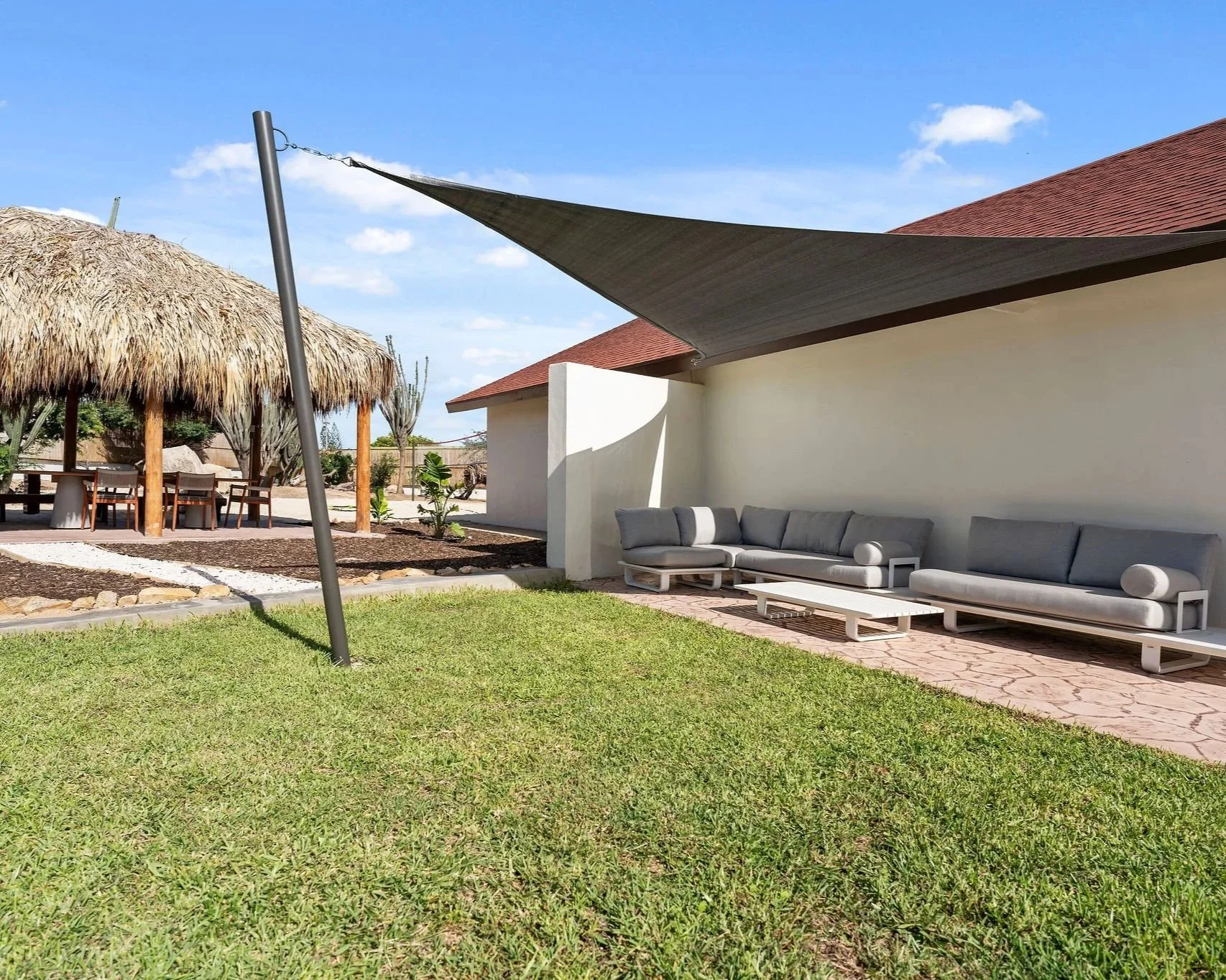 Outdoor patio area with modern gray sofas on a brick paved surface, shaded by a large triangular fabric sunshade, with a grassy lawn in the foreground and a thatched-roof hut with a dining table and chairs in the background under a clear blue sky.