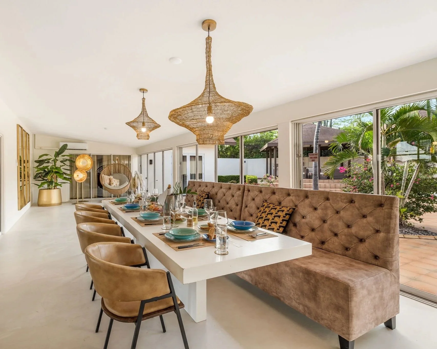 A modern dining room with a long white table set for a meal, surrounded by brown leather chairs and a tufted beige banquette. There are large windows with a view of tropical plants and a patio outside, and decorative woven pendant lights hang from the ceiling.