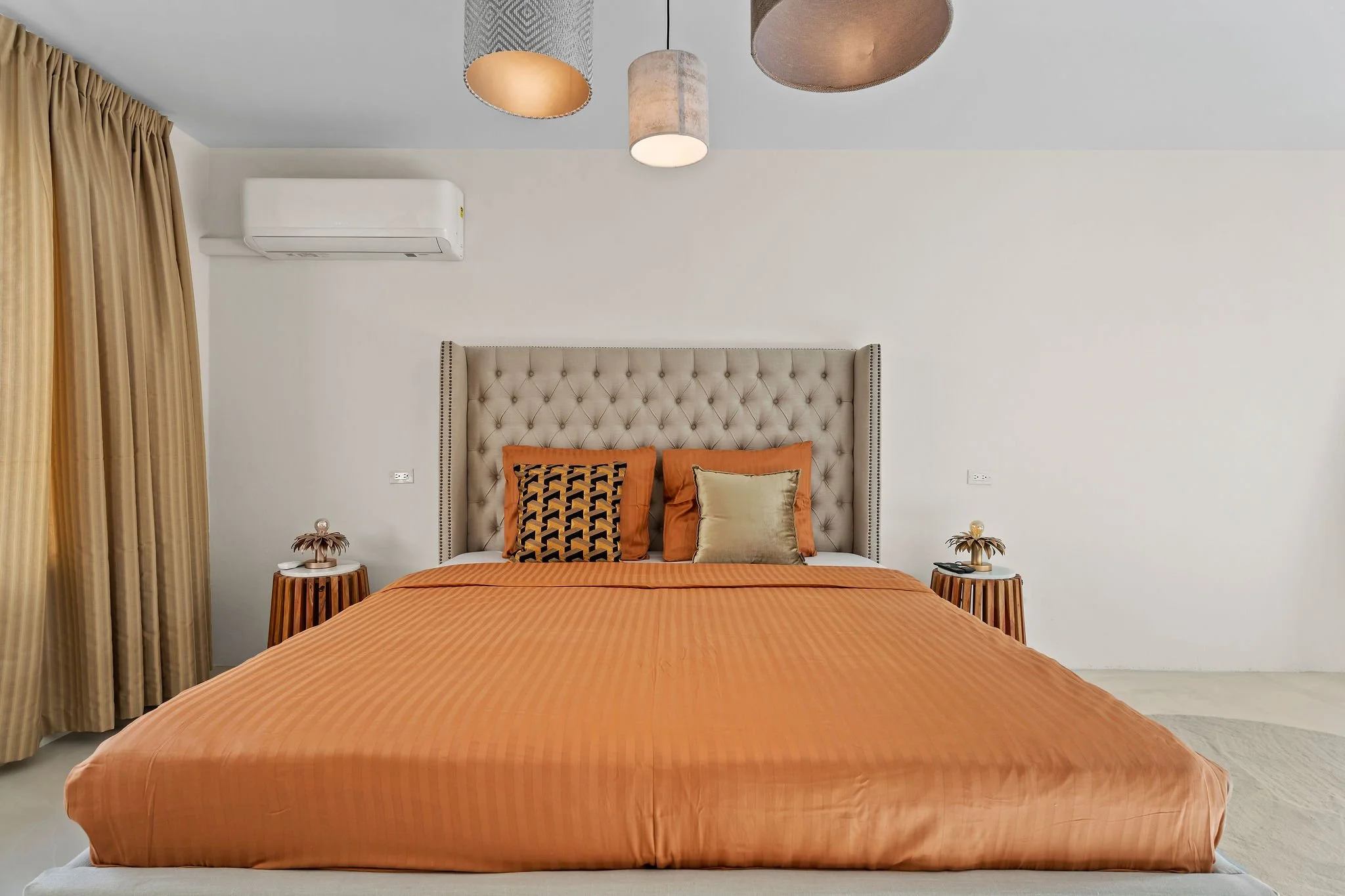 A bedroom with a bed covered in an orange bedspread, four pillows with orange, black and gold covers, a tufted beige headboard, two wooden nightstands with lamps, a white wall with a air conditioning unit and gold curtains on the left side.