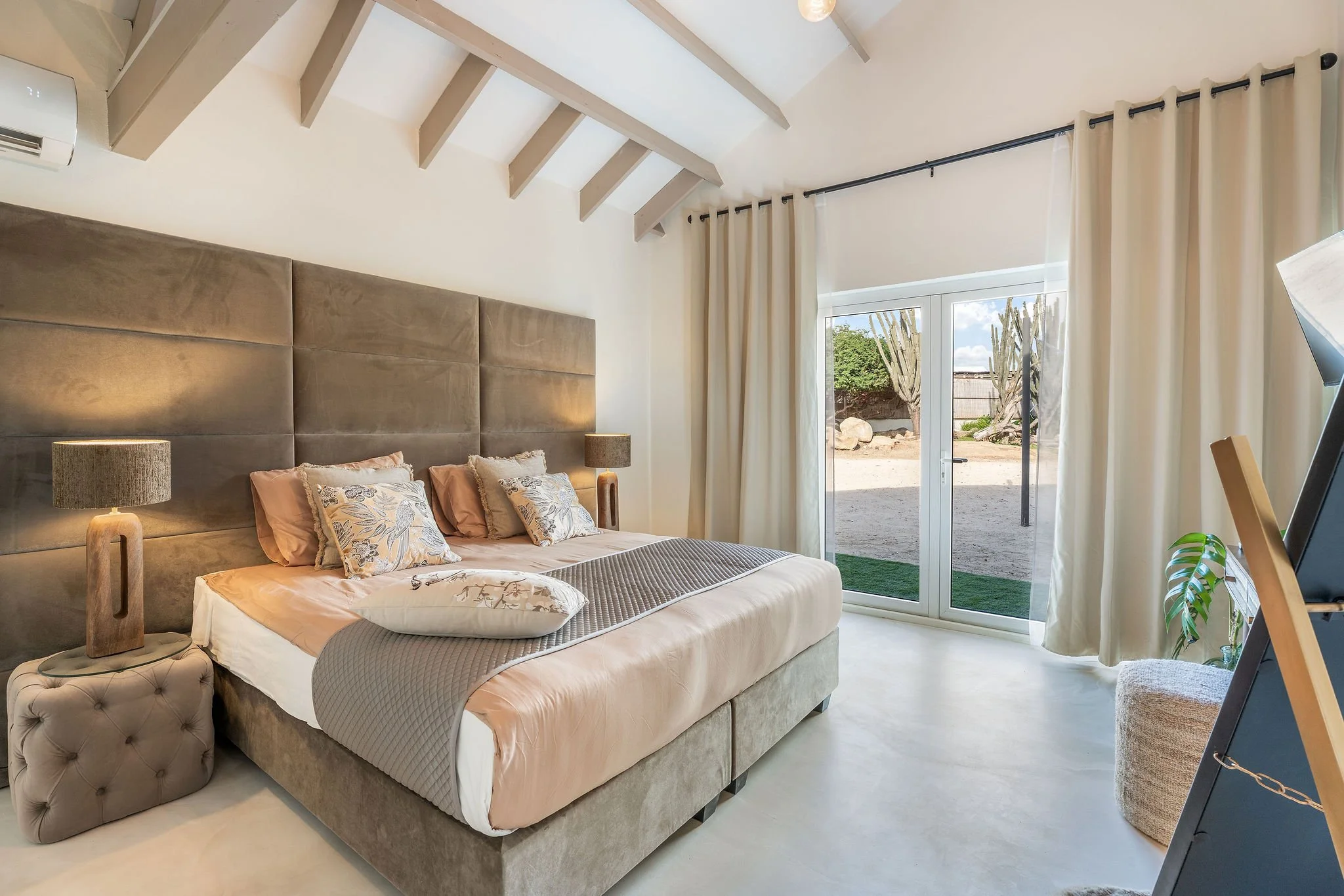 Modern bedroom with a large bed, beige upholstered headboard, and pillows with floral and neutral designs. Nightstands with lamps on either side of the bed. Large glass door with curtains leading outside, letting in natural light. Exposed beams on the ceiling and a small stool at the foot of the bed. Plants and other decorative elements in the room.