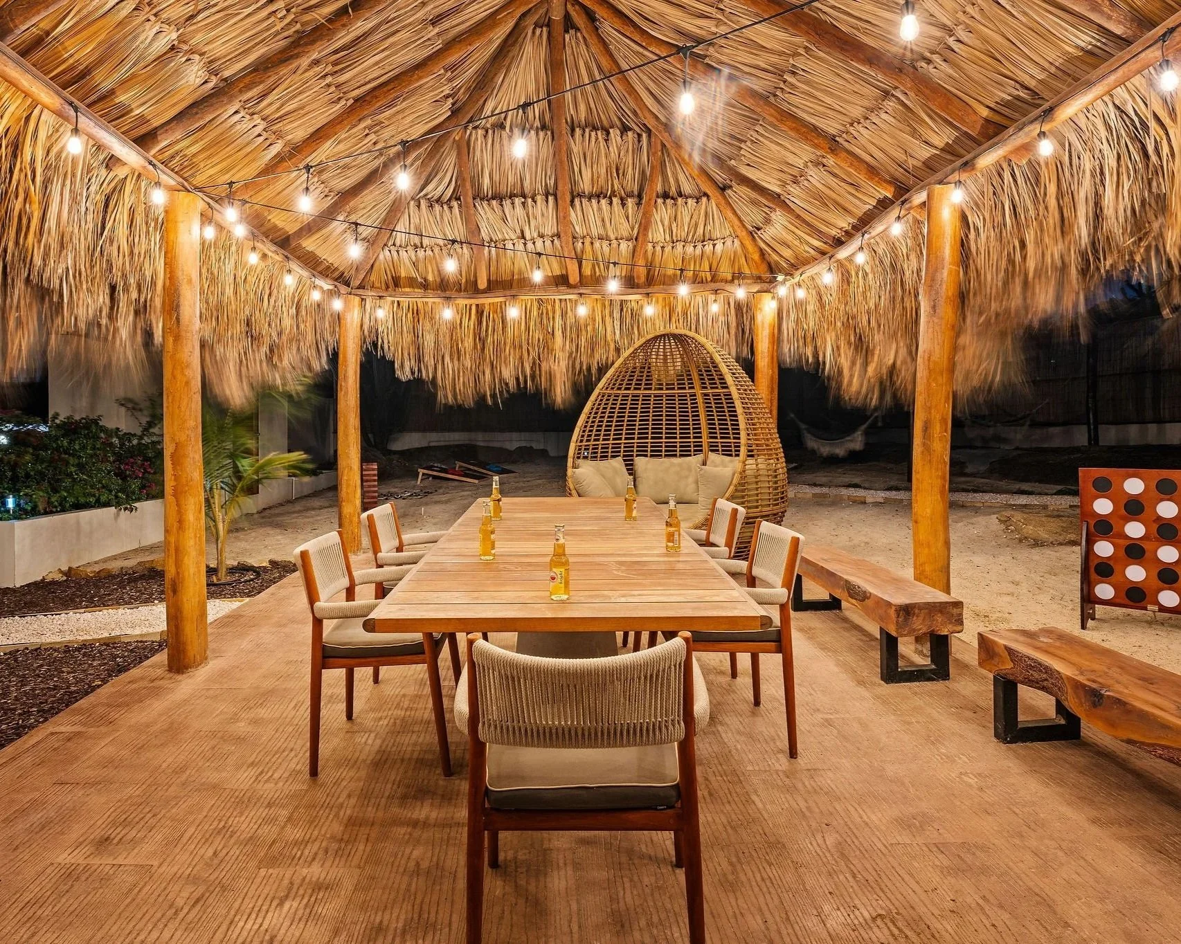 Outdoor dining area with a thatched roof, string lights, a long wooden table set with bottles, surrounded by chairs and benches, and a woven dome-shaped lounge in the background.