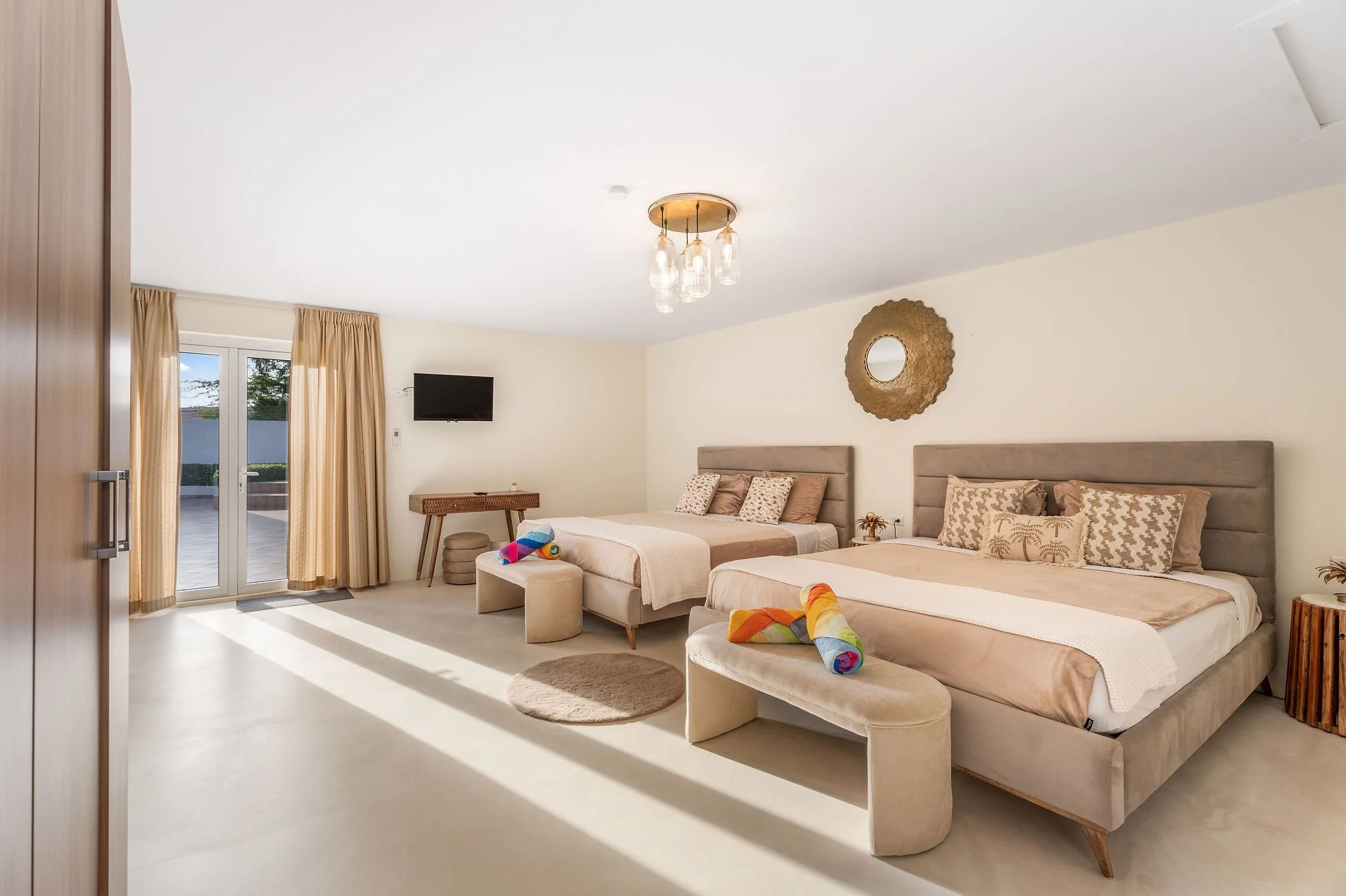 A hotel room with two beds, beige headboards, decorative pillows, and rolled towels on each bed. There's a small round rug in front of the beds, a wall mirror, a flat-screen TV, and a window with beige curtains allowing natural light into the room.
