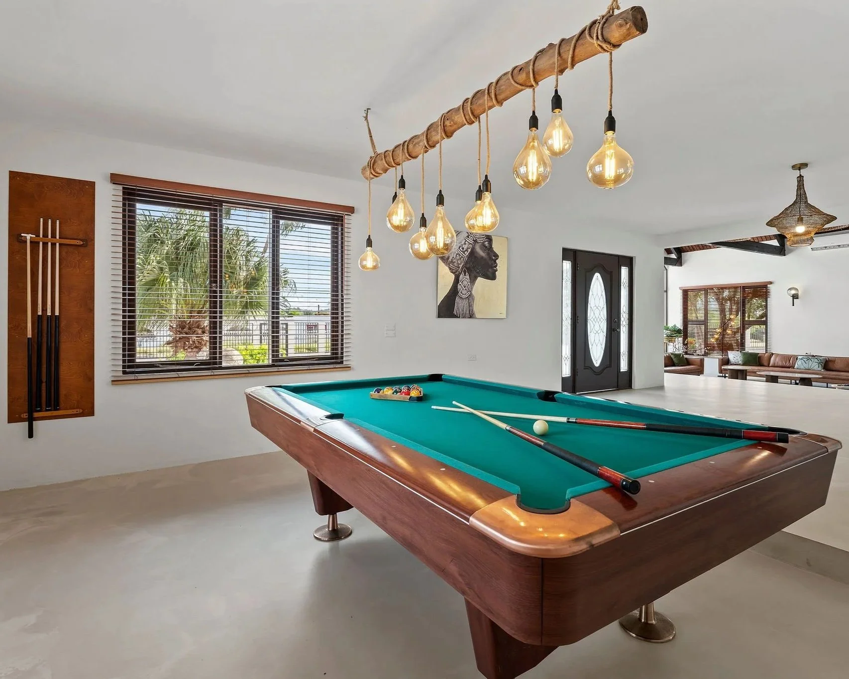 Indoor living room with a pool table, pool cues mounted on the wall, a painting of a woman in profile, large windows with blinds, and a door. Decorative lighting fixtures hanging from the ceiling.