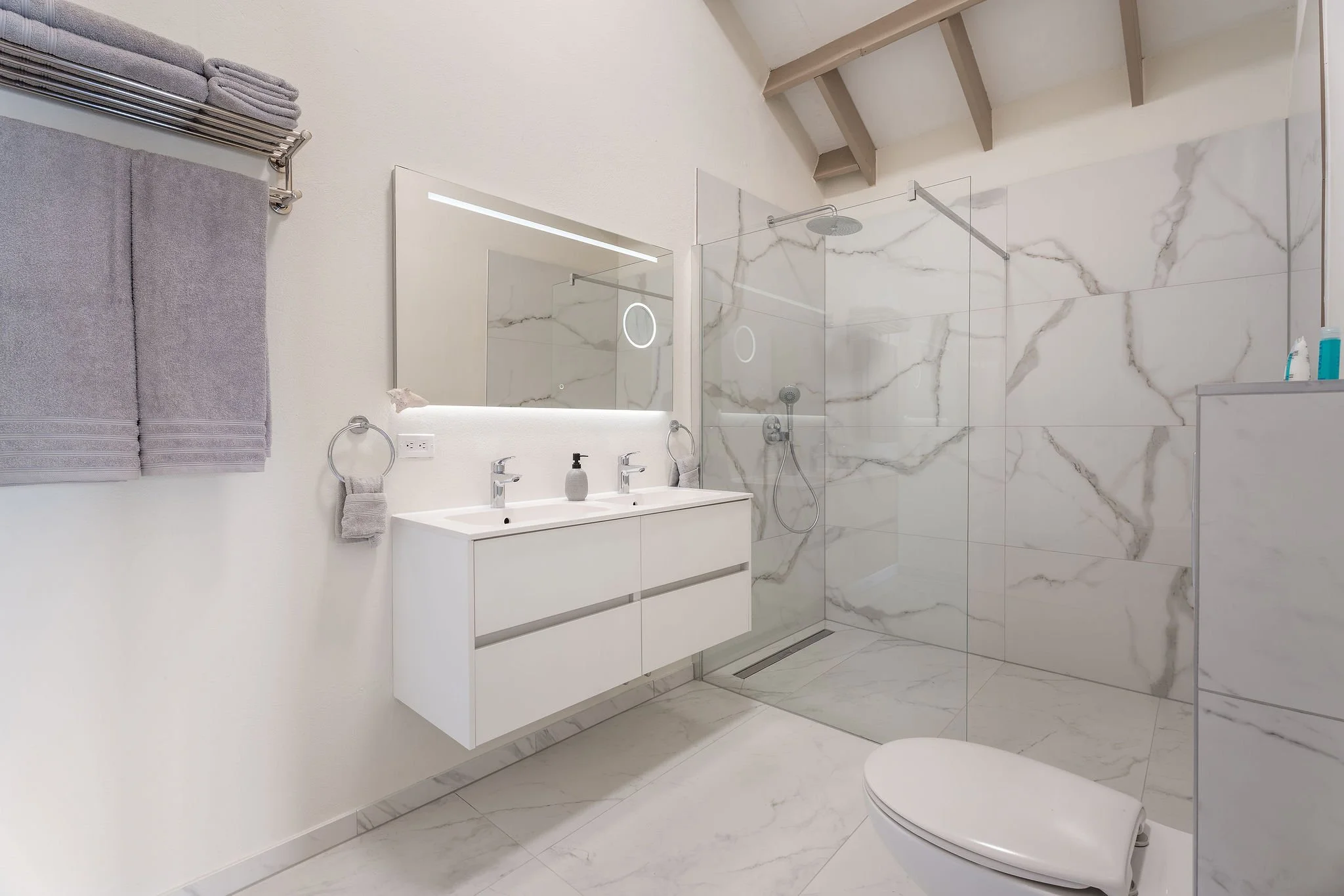 Modern bathroom with a marble shower wall, a white vanity with two sinks, gray towels on a heated towel rack, and a large mirror with integrated lighting.