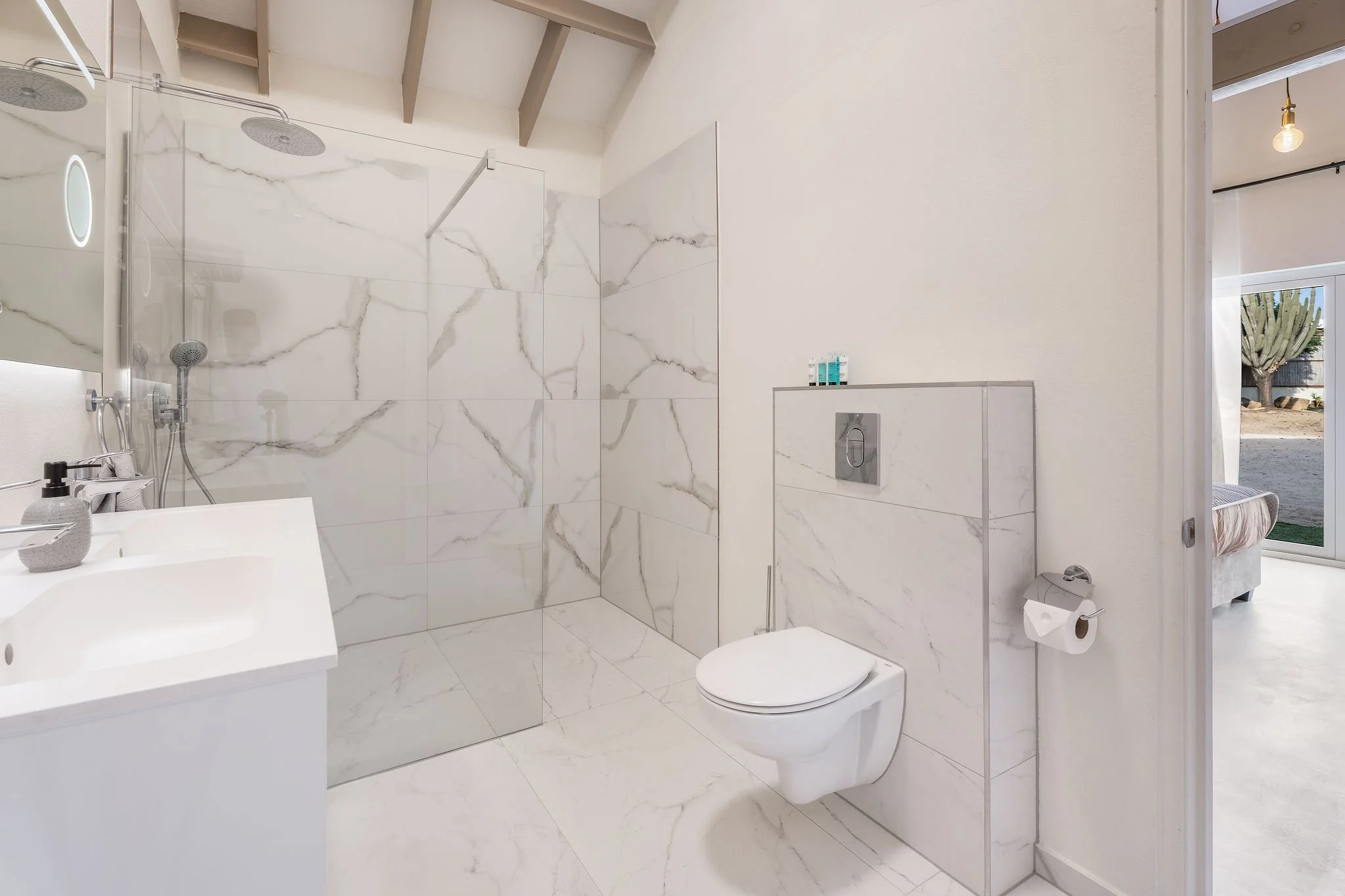A modern bathroom featuring a walk-in shower with marble tiles, a wall-mounted toilet, a small sink, and a window showing a cactus outside.