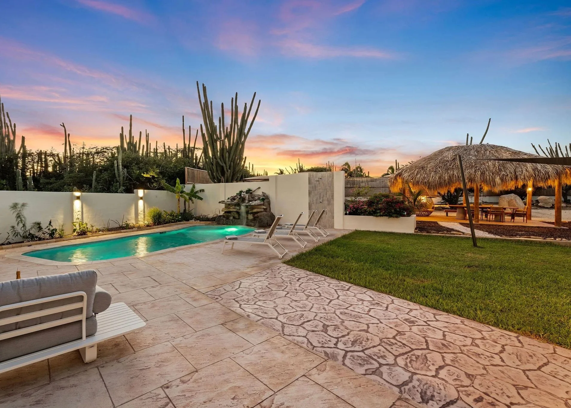 The villa casibari backyard with a swimming pool, lounge chairs, a thatched roof tiki hut with seating, desert plants including tall cacti, and a sunset sky with pink, purple, and orange hues.