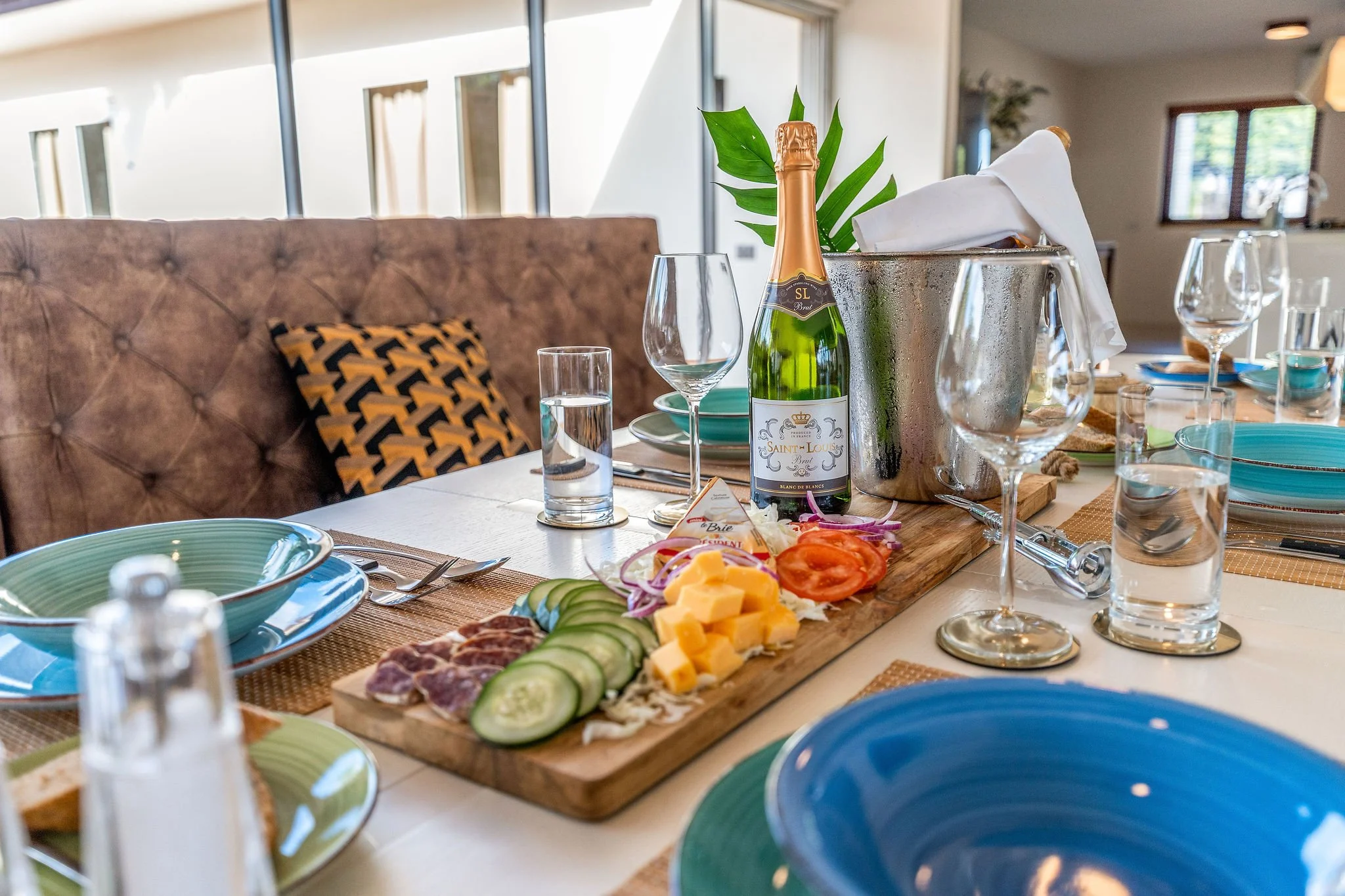 A dining table set for a meal with plates, glasses, and cutlery, featuring a charcuterie board with sliced cucumbers, cheese, and meats, a bottle of sparkling wine, and a metal ice bucket with a napkin, in a well-lit room with large windows and a cushioned bench with a decorative pillow.