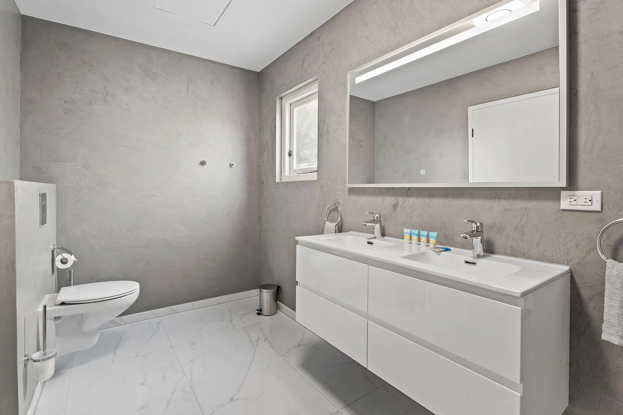 Modern minimalist bathroom with double sink vanity, large wall mirror, small window, and toilet, all in neutral tones.