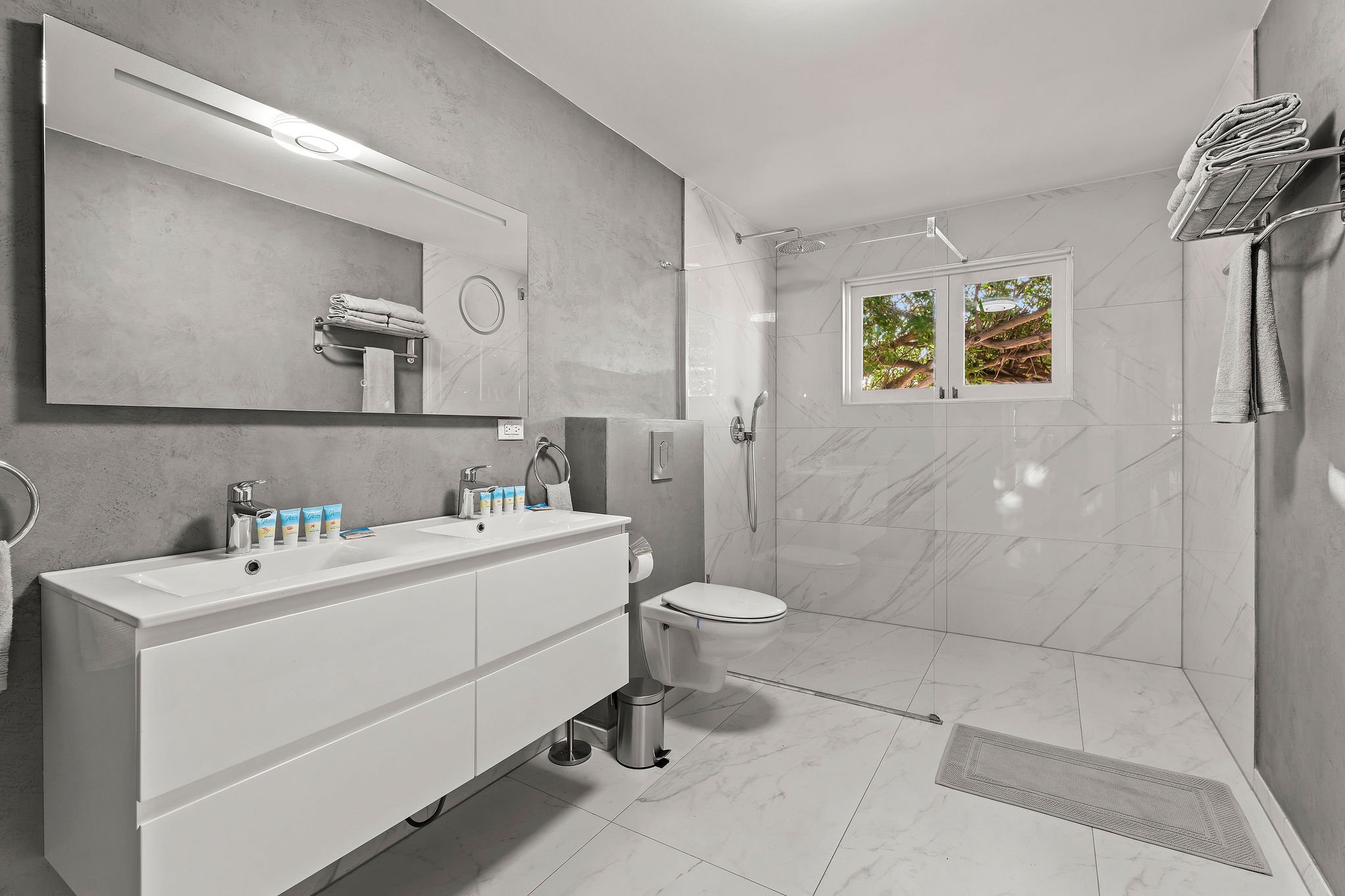 Modern bathroom with white vanity, rectangular mirror, wall-mounted faucet, toiletries, toilet, walk-in shower with glass door, large window showing greenery outside, shelves with towels, and gray floor mats.