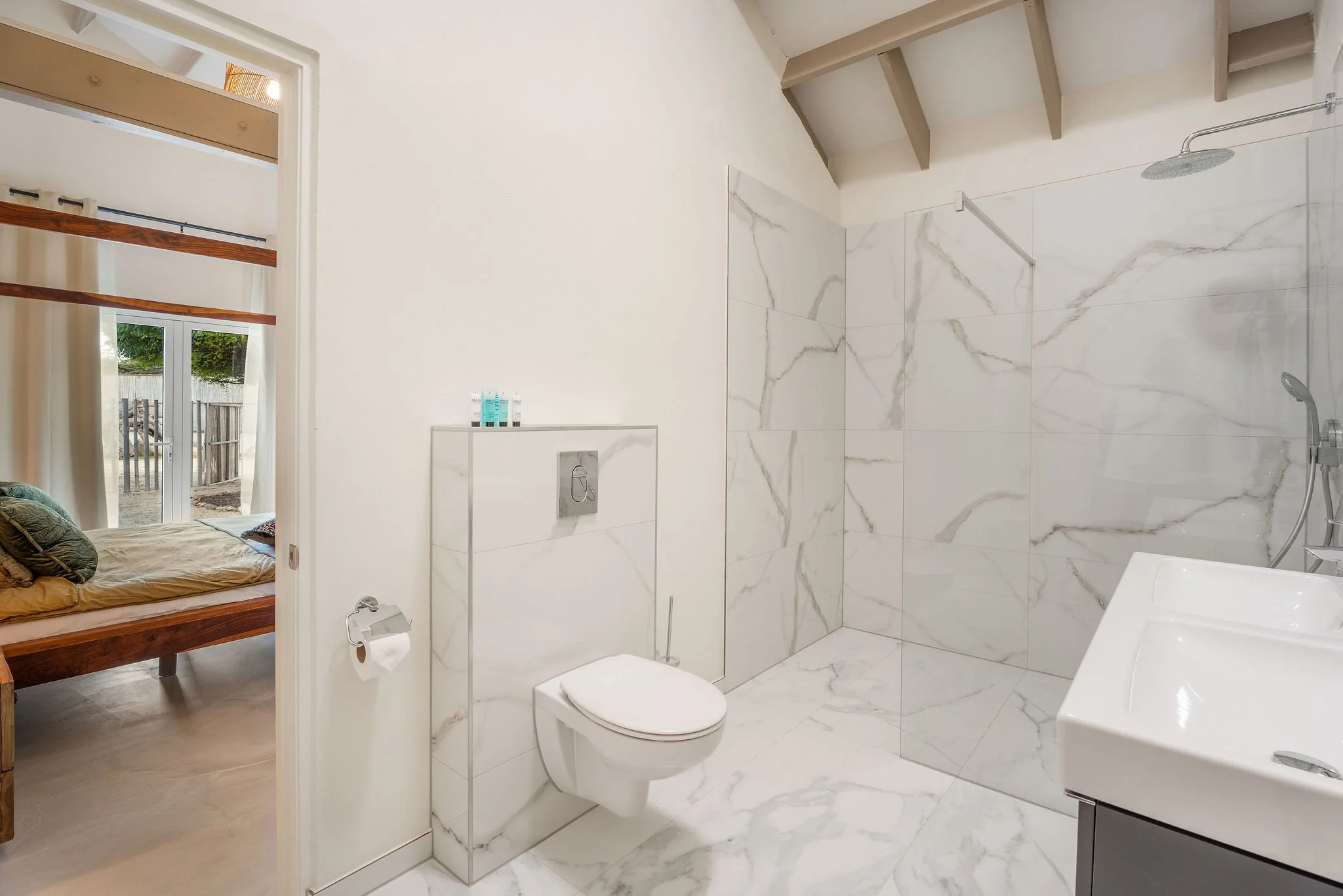 A modern bathroom with white marble walls and flooring, a walk-in shower with a rain showerhead, a toilet, and a small sink. The bathroom is adjacent to a bedroom with a window and wooden shelves.