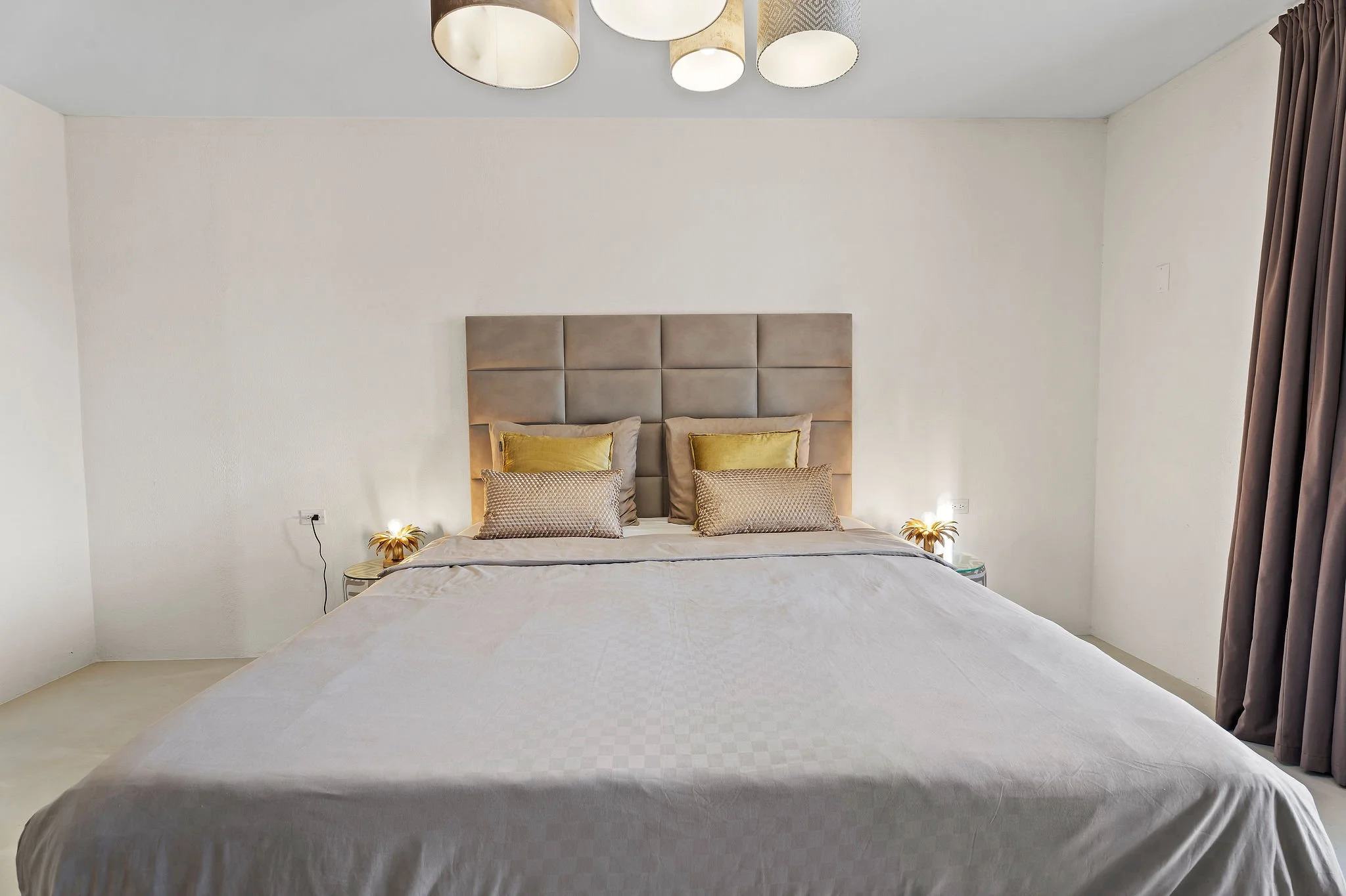 A neatly made bed with a padded headboard, multiple pillows in shades of beige and gold, side tables with decorative lamps, and curtains on the right side of the window, all in a modern minimalist bedroom with light-colored walls and ceiling.