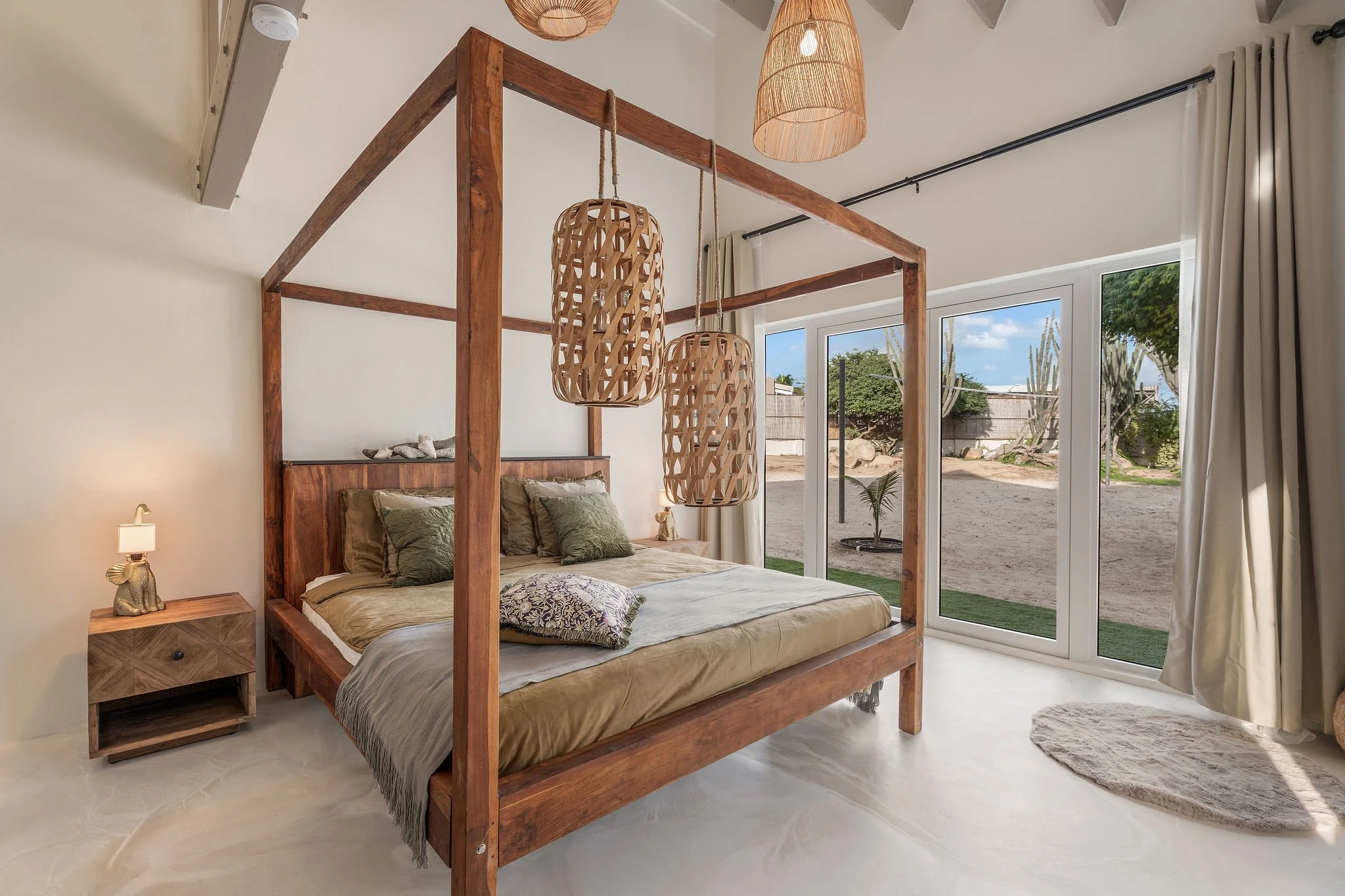 A bedroom with a wooden four-poster bed, bedding, pillows, two hanging pendant lights, a nightstand with a decorative lamp, and large glass doors revealing an outdoor view with trees and a sandy area.