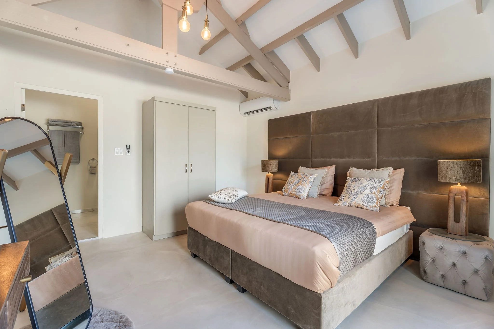 Interior of a bedroom with a large bed featuring a beige bedspread and decorative cushions, bedside tables with lamps, a white wardrobe, a mirror, and exposed ceiling beams with hanging light bulbs.