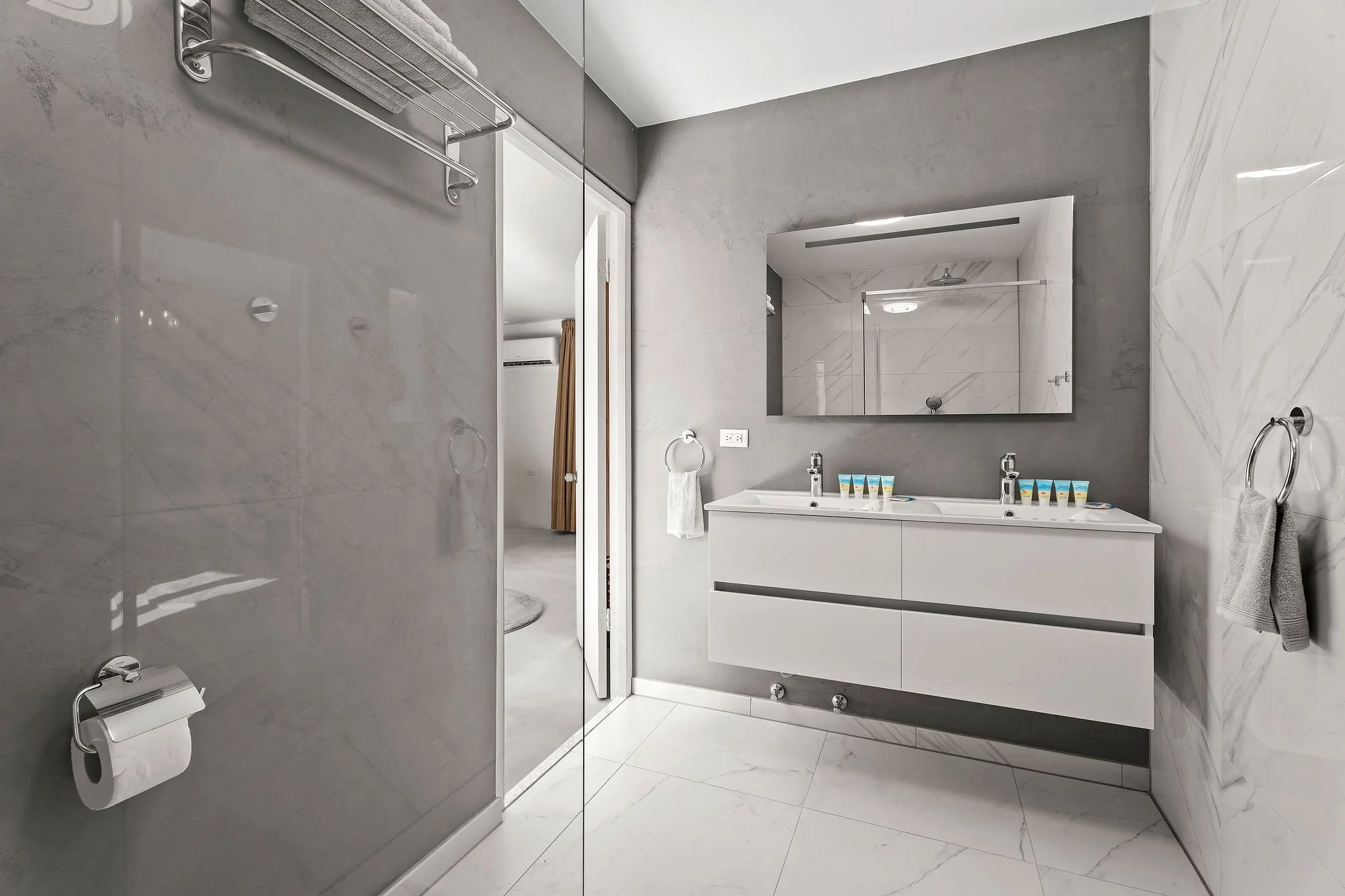 Modern bathroom with a large mirror, double sink vanity, gray and white marble walls, and a towel rack with folded towels.