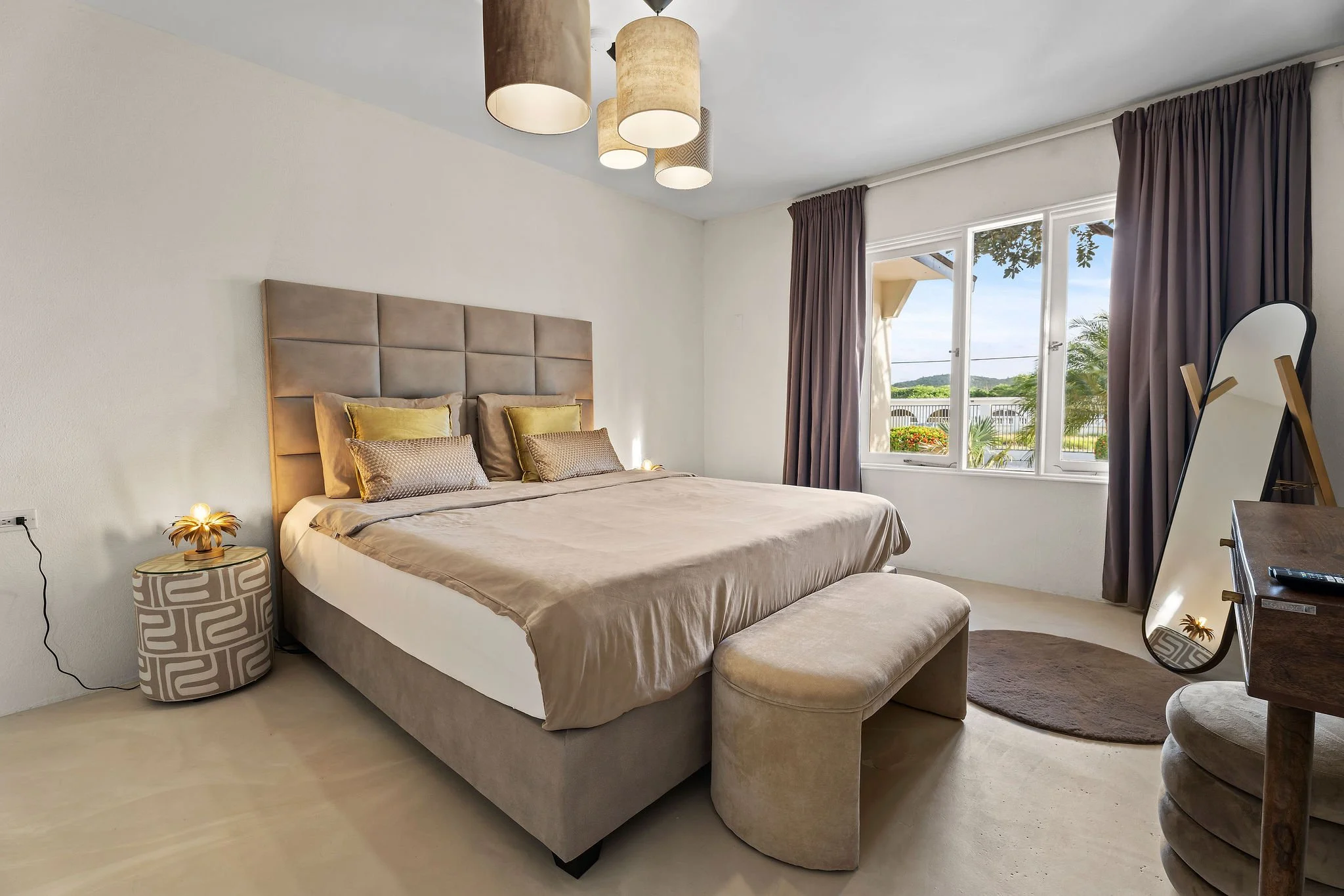 A bedroom with a large bed that has beige and gold pillows, a padded headboard, and a matching beige bench at the foot of the bed. There is a small round nightstand with a decorative lamp shaped like a flower next to the bed. To the right, a large window with dark curtains shows a view of greenery and a balcony outside. An oval mirror and a wooden dresser are also visible.