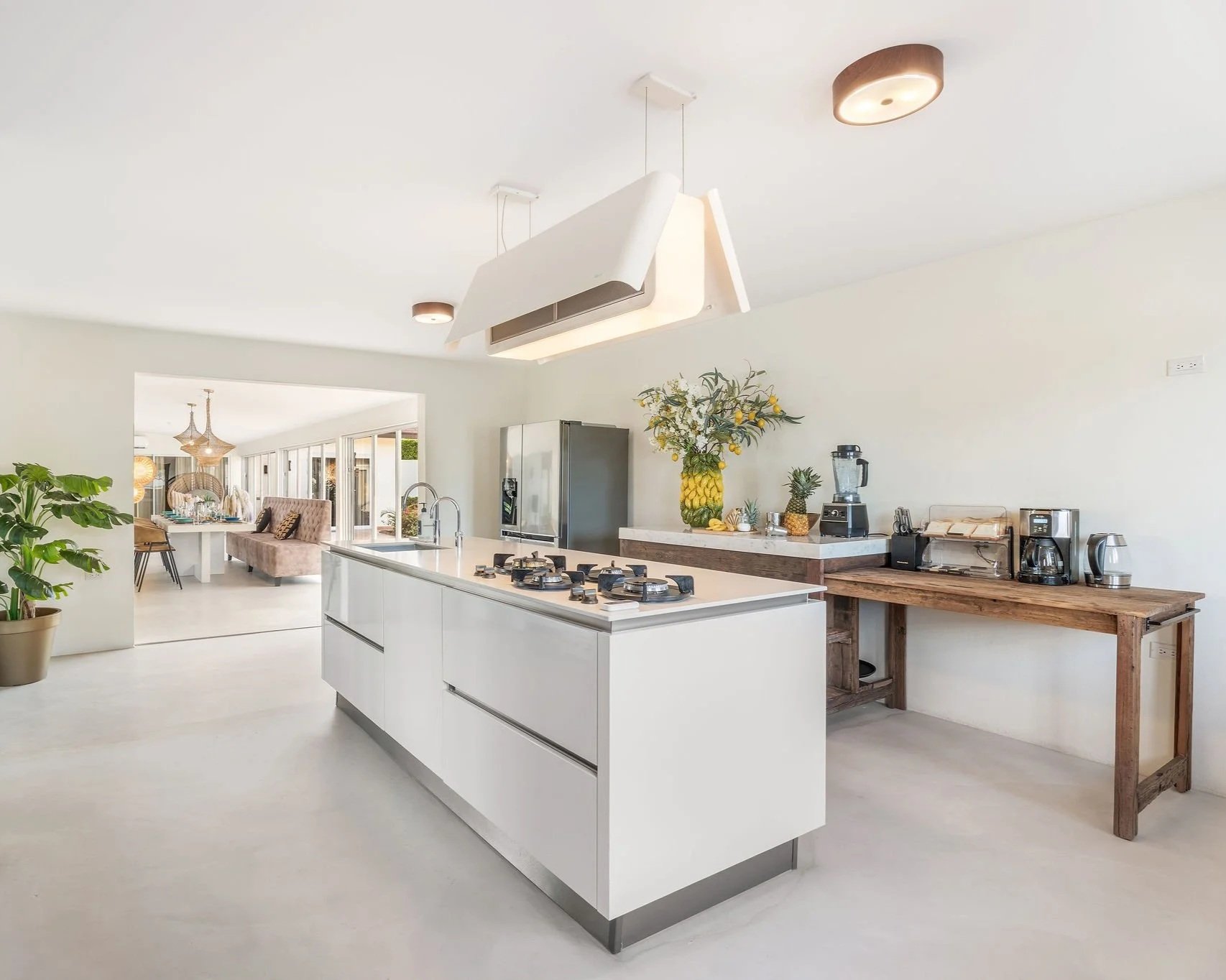 Modern kitchen with white island and stainless steel appliances, wooden countertop with coffee maker and blender, decorative flowers, and an adjacent dining area with chandelier and seating.