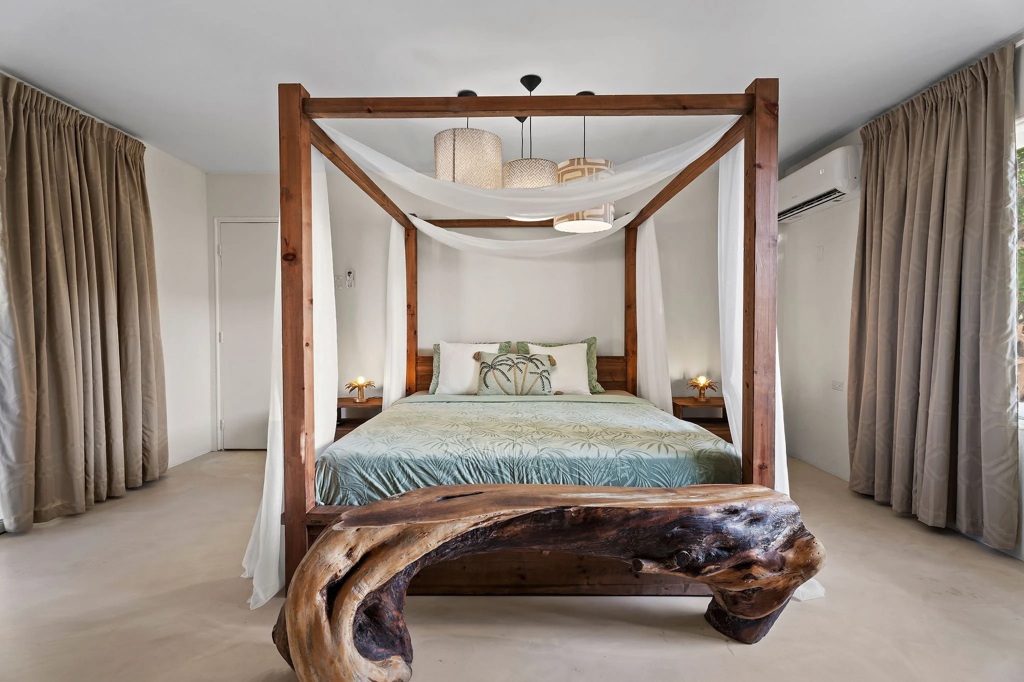 A bedroom featuring a four-poster bed with a rustic wooden frame and a gnarled wooden base. The bed has tropical-themed pillows and a light-colored bedspread. Two bedside lamps with golden-toned bases are on small tables on either side of the bed. The room has beige curtains on the windows and an air conditioning unit on the right wall.