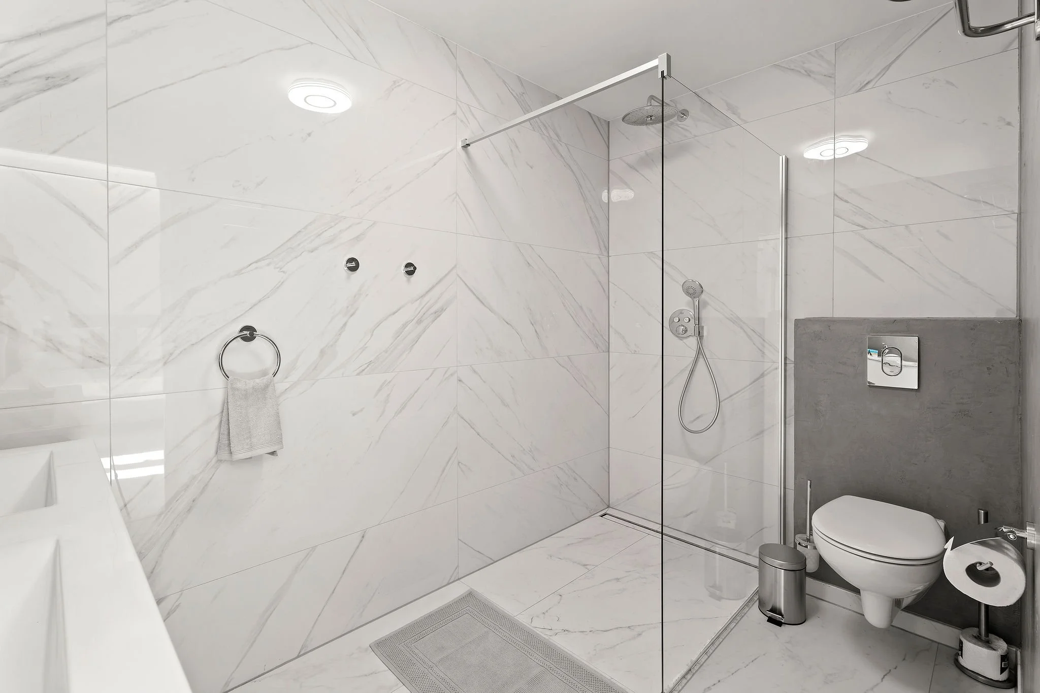 Modern bathroom with white marble walls, a toilet, a walk-in shower with a glass door, a rain shower head, and a handheld shower spray. There is a towel on a ring hook, a small trash bin, and a toilet paper roll on top of a stand.