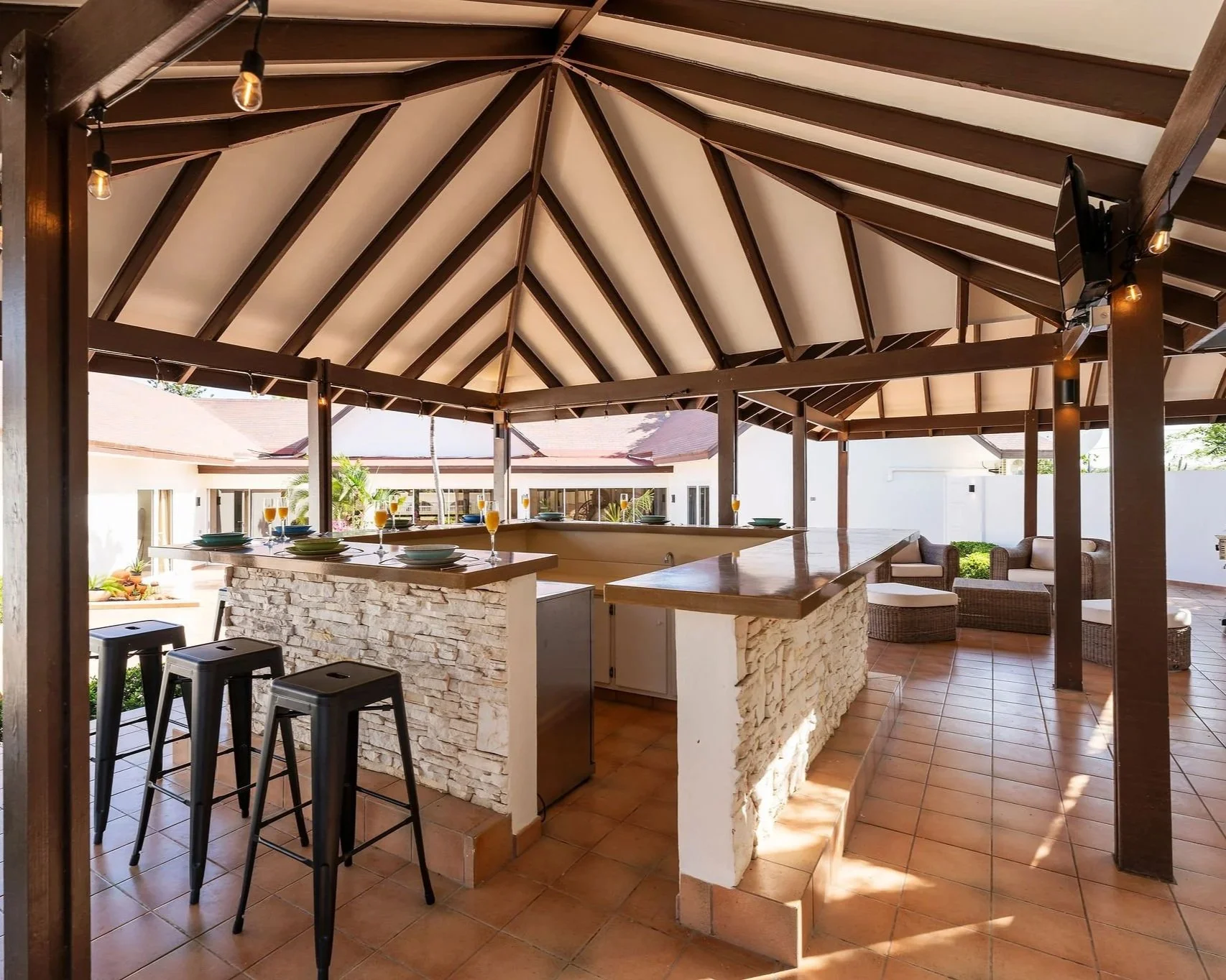 Outdoor covered patio area with a high peaked wooden ceiling, string lights, a bar with stone facade, black bar stools, and patio furniture in the background.