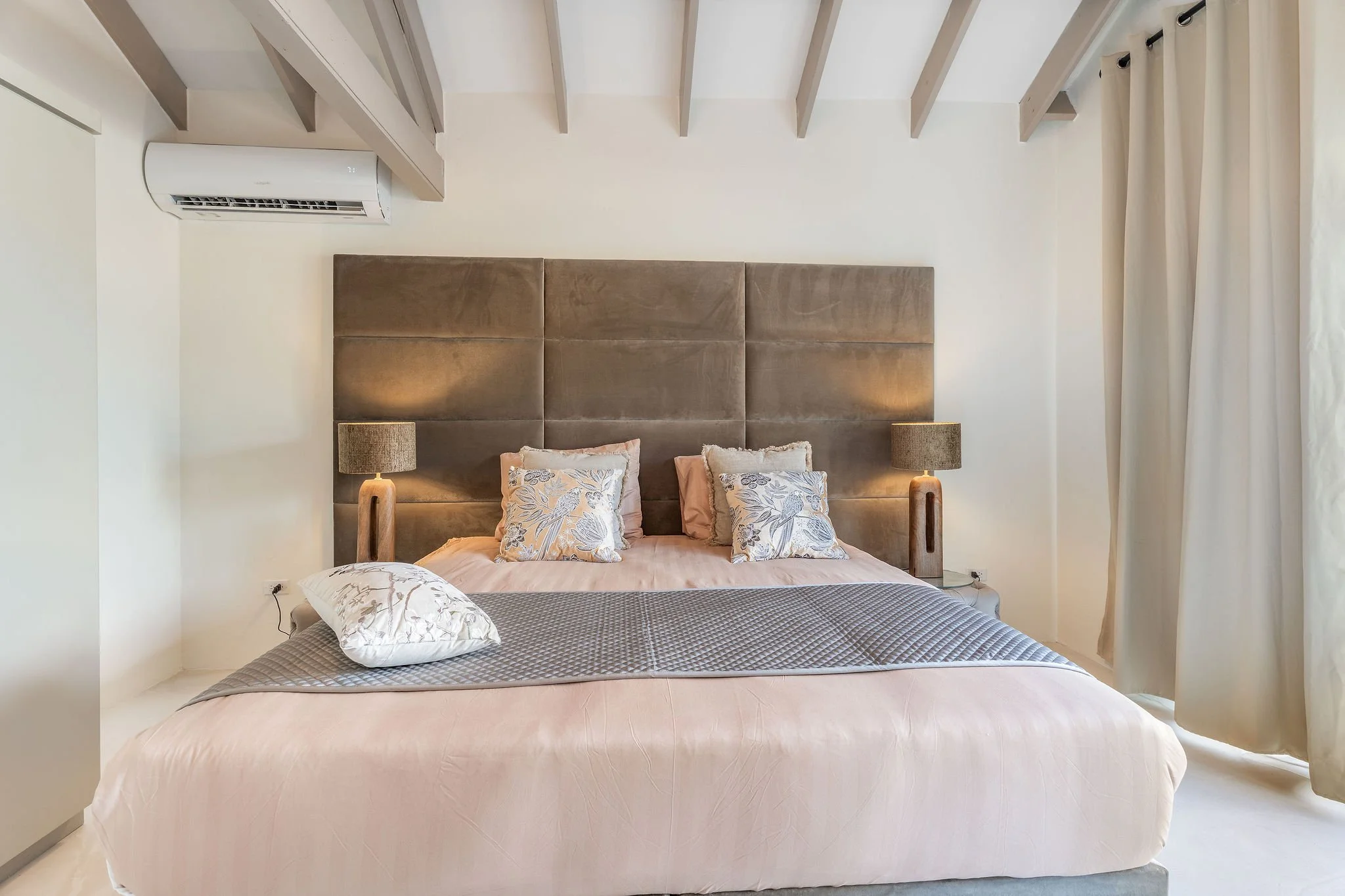 A bedroom with a large bed featuring a padded headboard, multiple decorative pillows, two bedside lamps, and an air conditioning unit on the wall.