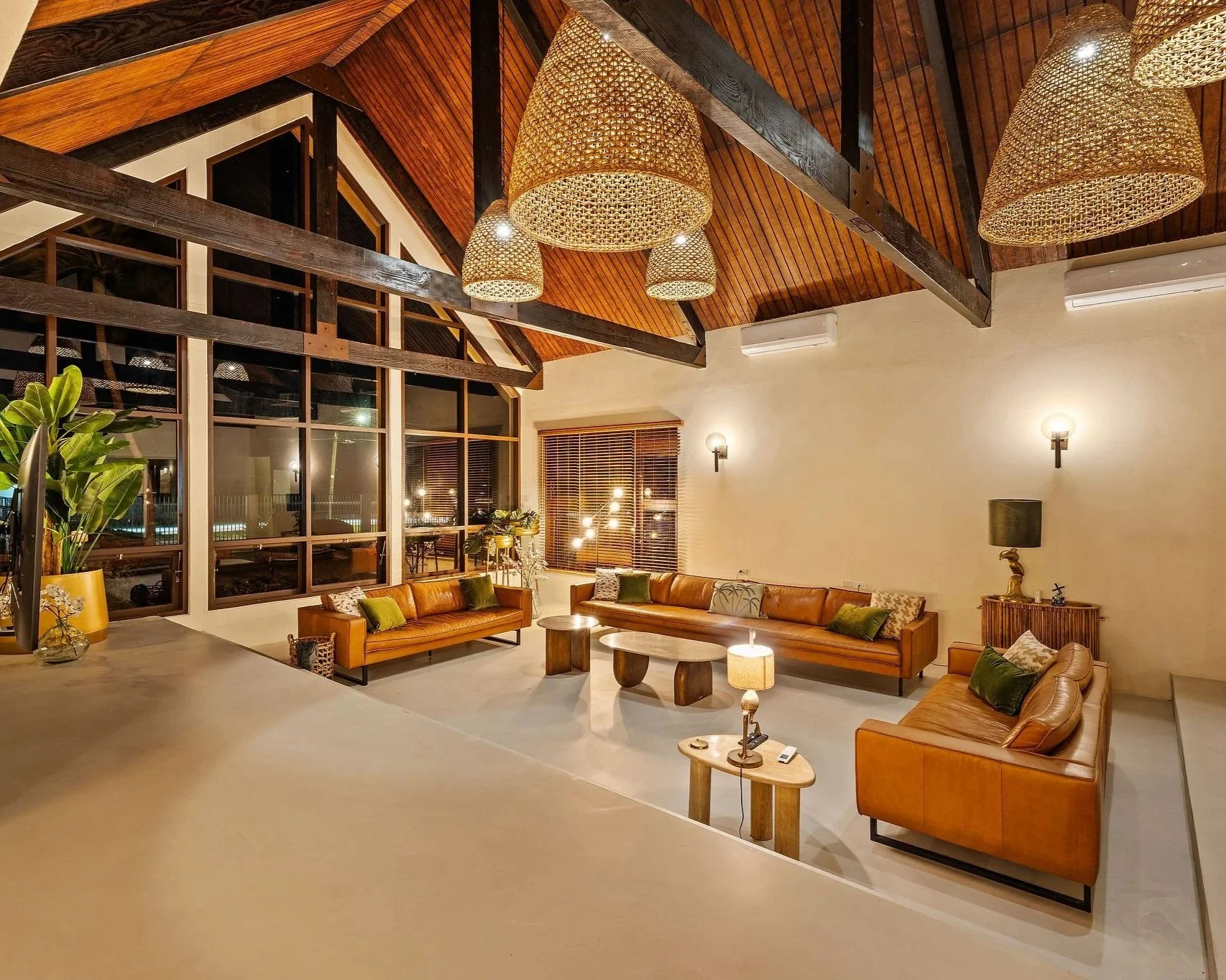 A spacious living room with high vaulted wooden ceilings, multiple large woven pendant lights, and large windows with wooden blinds. There are three leather sofas with pillows, a few coffee tables, a small side table with a lamp, and some indoor plants.