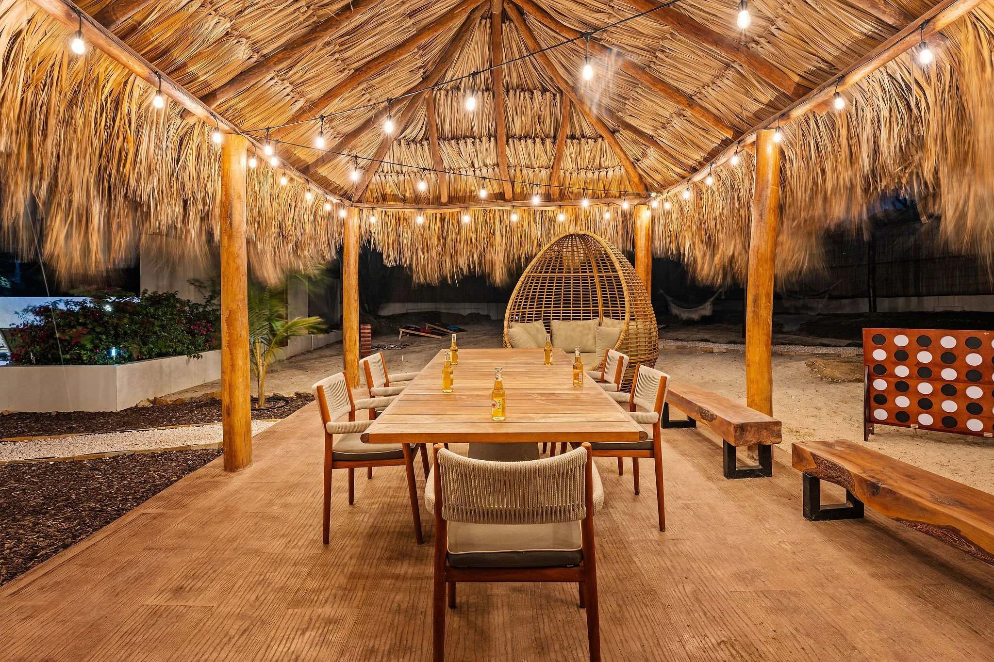 Villa casibari dining area under a thatched roof with string lights, a wooden table, cream chairs, a wicker pod chair, and some beer bottles on the table.