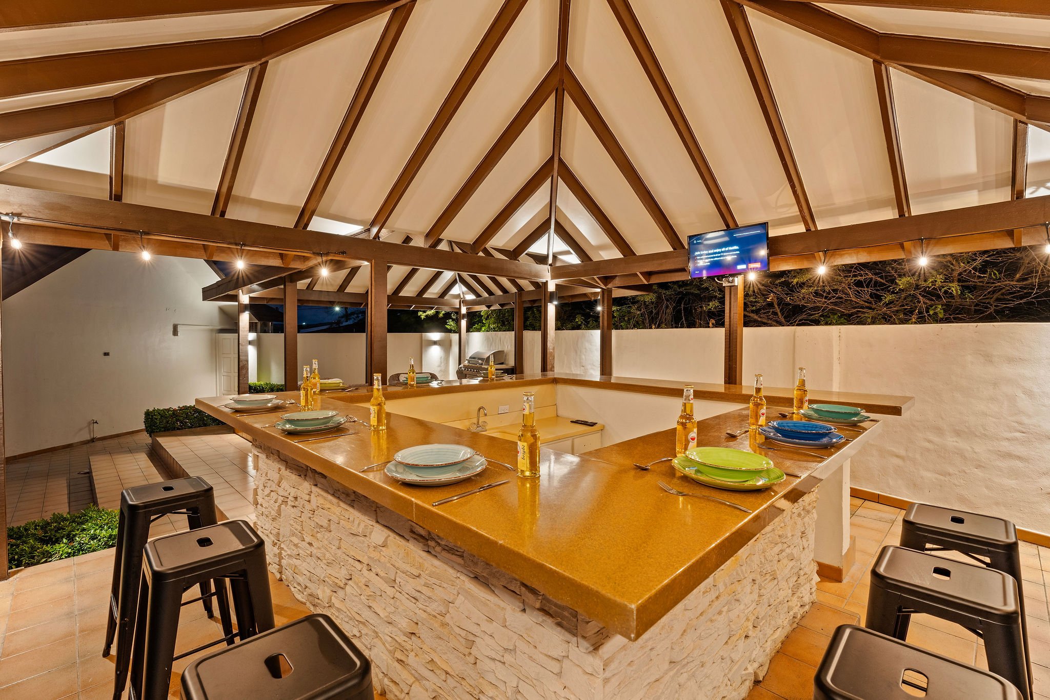 Outdoor patio area with a large wooden and stone counter, set for a meal with colorful plates, bottles, and utensils. A roof with exposed wooden beams and string lights. A nearby window, a TV mounted on the beam, and some chairs and greenery visible.