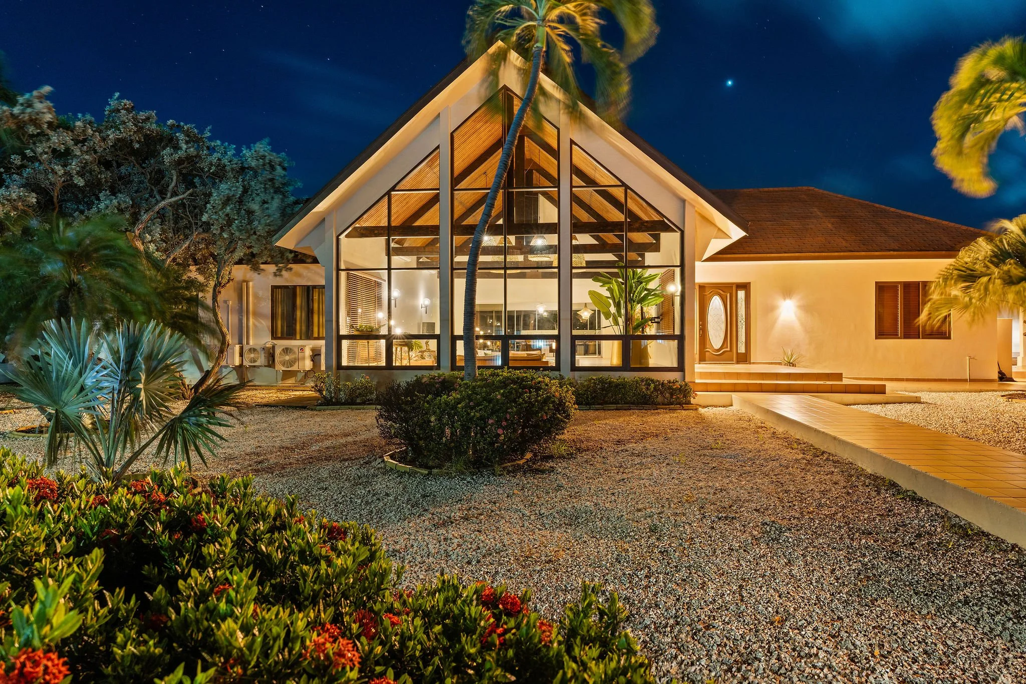 casibari villa with large glass windows and warm interior lighting at night, surrounded by tropical plants and trees, with a gravel pathway leading to the entrance.