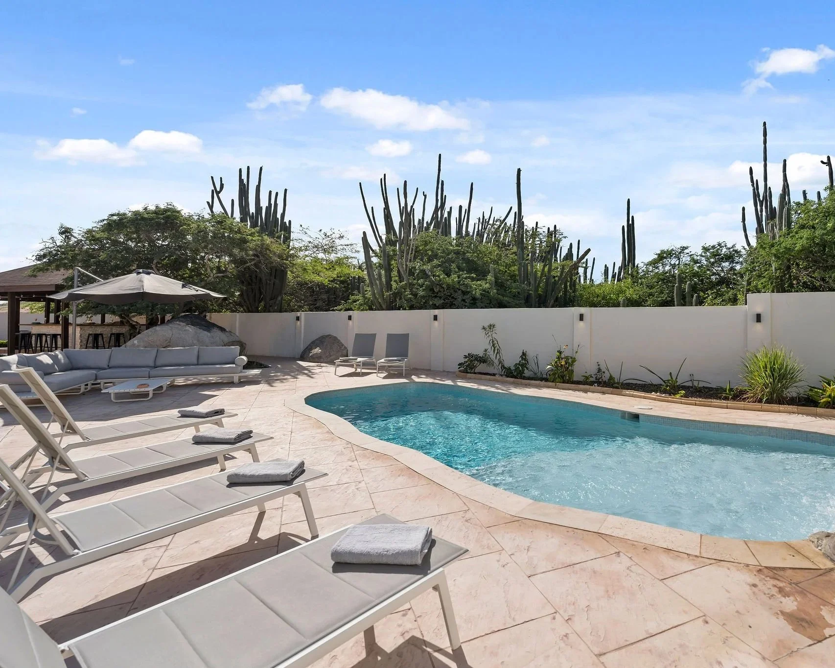 A backyard with a swimming pool, lounge chairs, a large gray outdoor sofa, patio umbrella, and desert cactus plants in the background under a blue sky with scattered clouds.