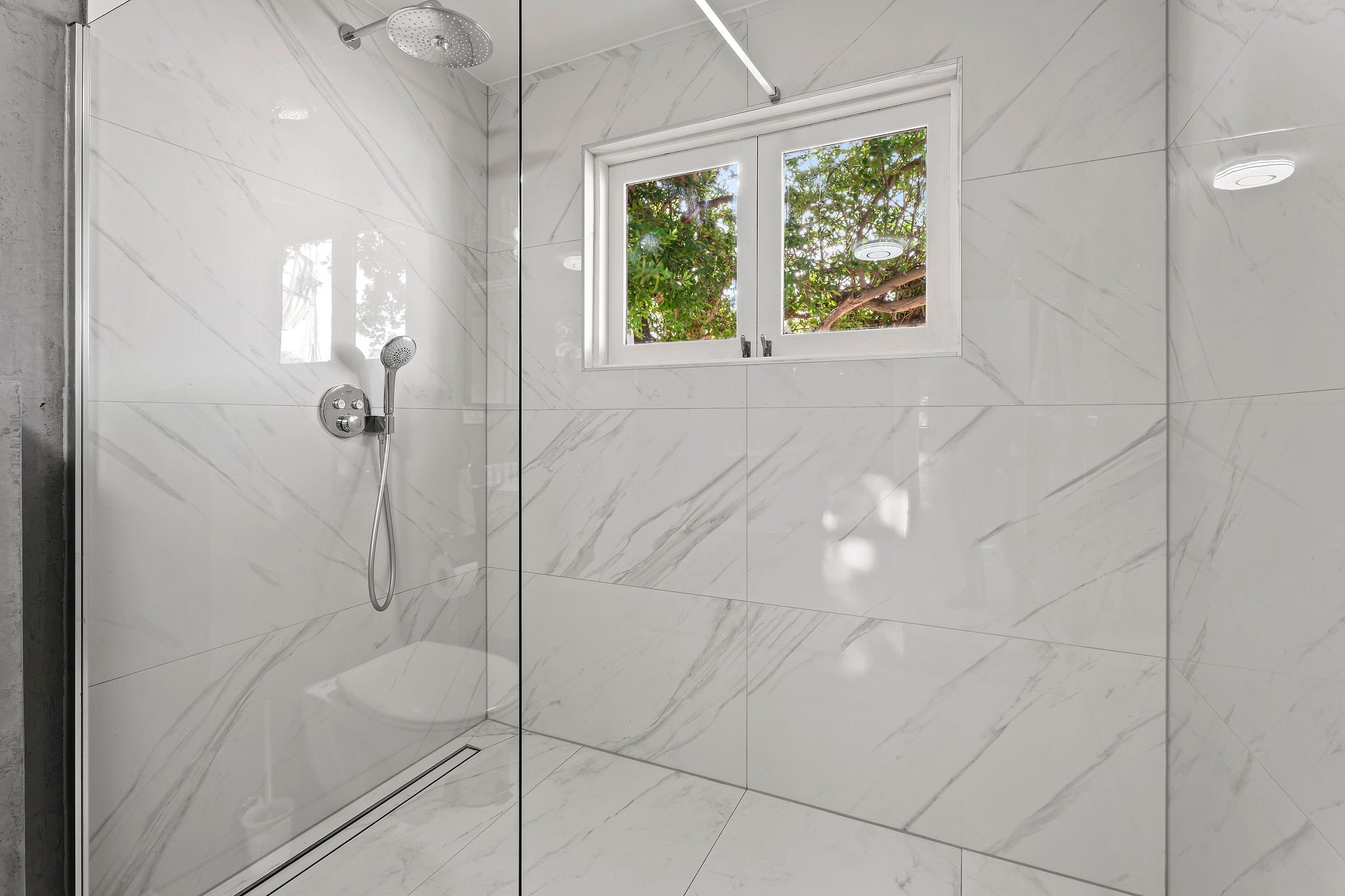 Modern bathroom shower area with marble tiles, a ceiling-mounted shower head, a handheld shower, and a window showing green foliage outside.