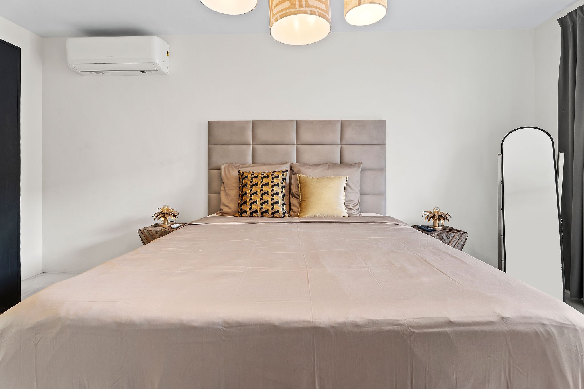 A neatly made bed with a beige headboard, decorated with three pillows in beige, black and gold, and yellow. The room has white walls, a ceiling light fixture with three beige lampshades, dark curtains on the right, and a white air conditioning unit on the upper left wall.