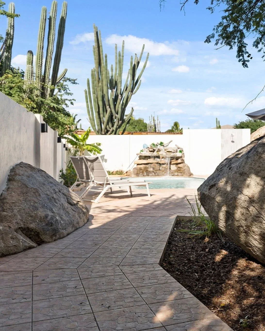 A backyard patio with a swimming pool, cacti, and large rocks, under a blue sky with clouds.