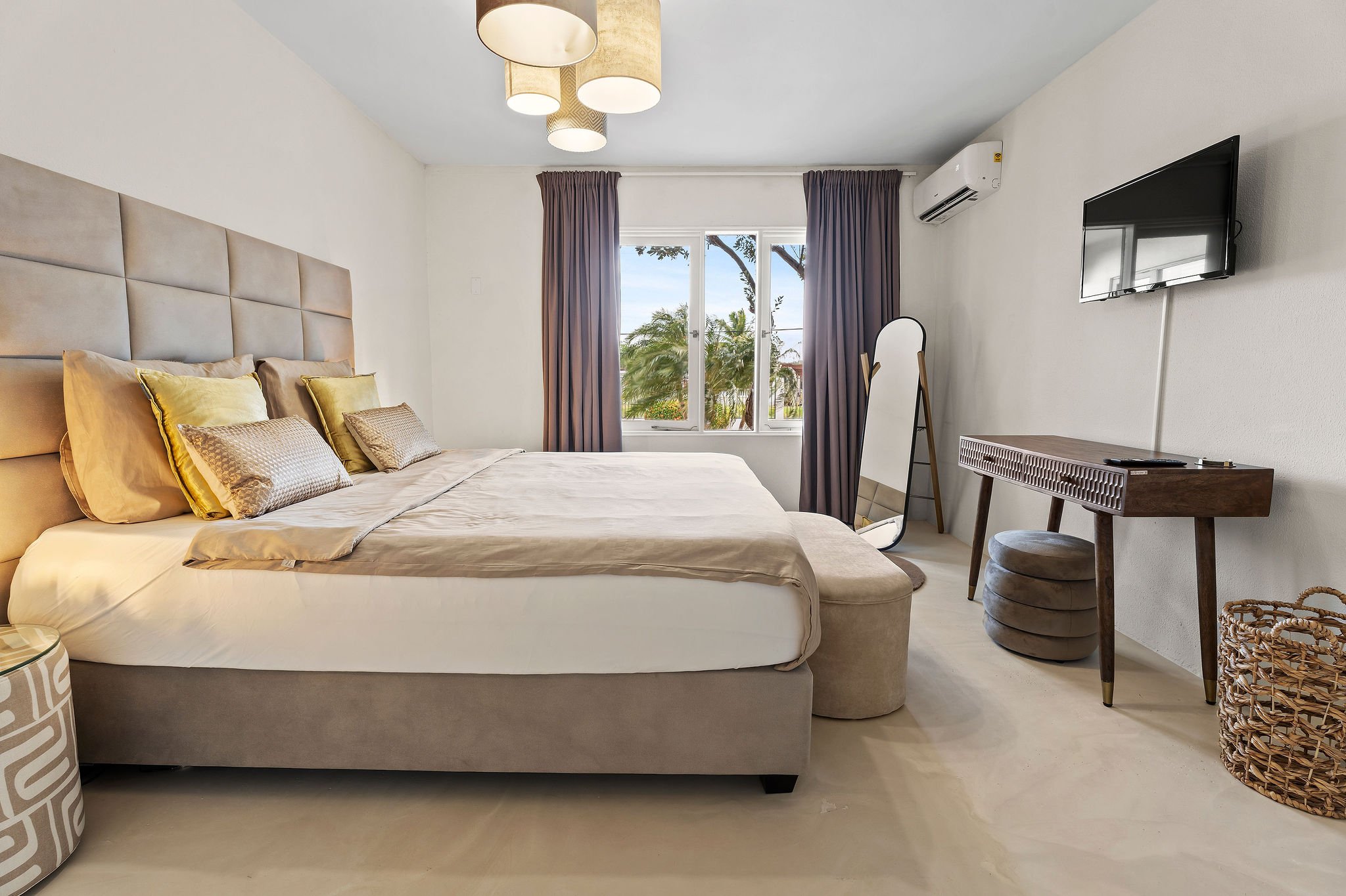 A bedroom with a large bed, multiple pillows, a wardrobe, a TV mounted on the wall, a mirror, and a large window with curtains showing a view of palm trees and sky.