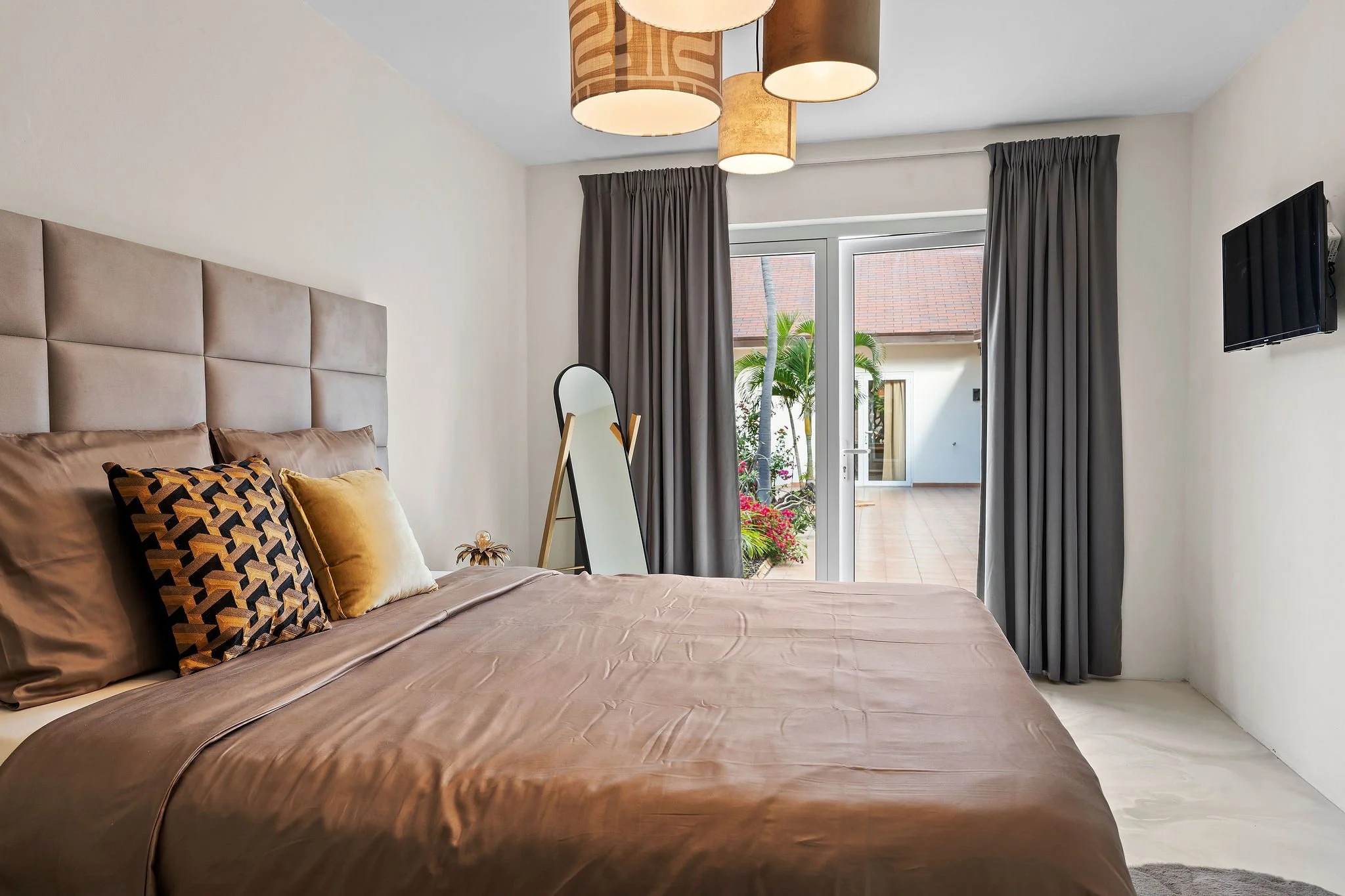 A bedroom with a large bed covered in brown bedding and decorated with multiple pillows. There are dark curtains over a glass door that opens to an outdoor patio with plants. A standing mirror is placed near the door, and a wall-mounted television is visible on the right side.