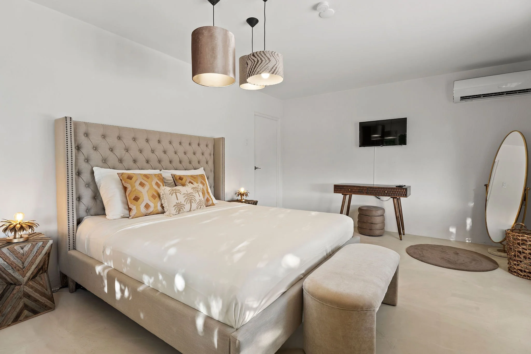 A modern bedroom with a beige tufted headboard, white bedding, and decorative pillows featuring geometric and palm leaf patterns. There are two bedside tables with gold lamps, a wall-mounted TV, a wooden console, a standing mirror with a gold frame, and a beige ottoman. The room has white walls, a white ceiling with three pendant lights, and an air conditioning unit.