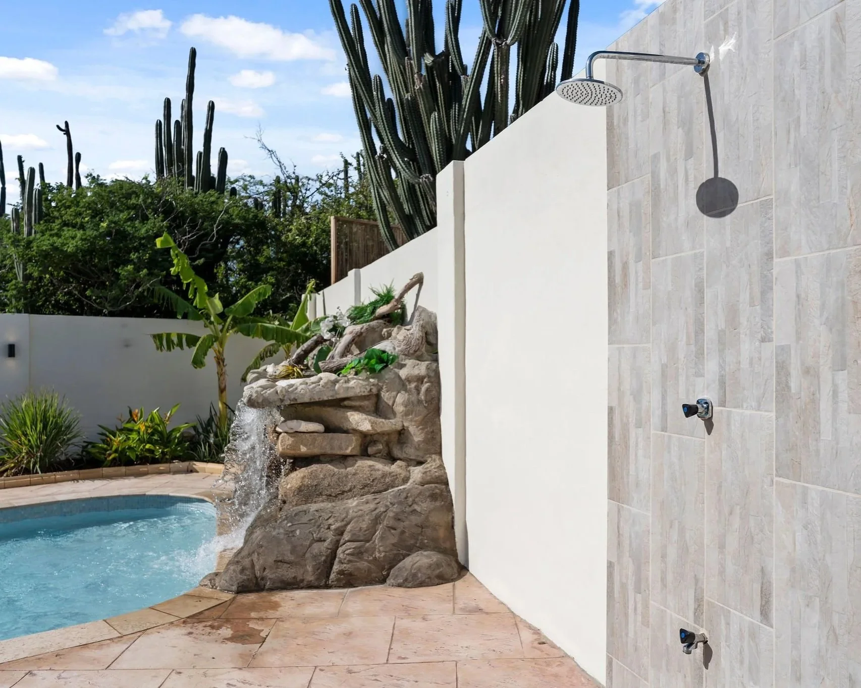 Outdoor pool with rock waterfall feature, surrounded by tropical plants and cacti, with a shower area on the tiled deck.