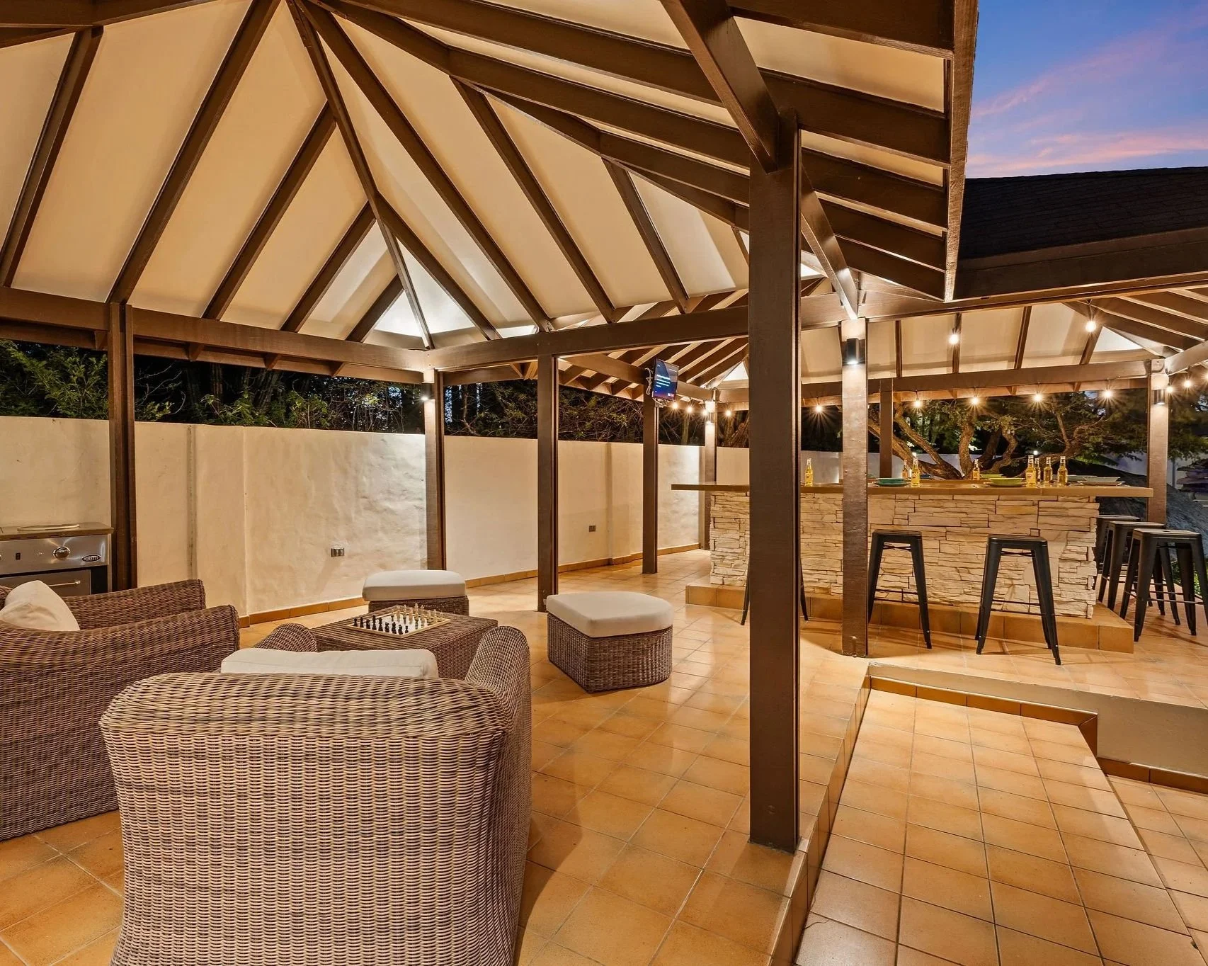 An outdoor bar area with wicker seating, a stone-clad bar counter with beer bottles, bar stools, a roof with exposed wooden beams, and string lights as evening sky is visible.