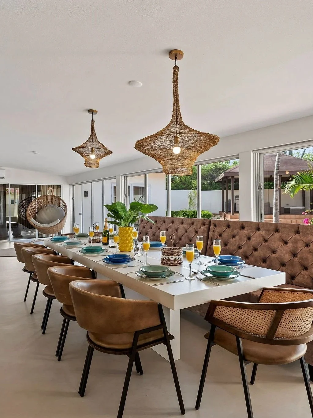 A modern dining room with a long white table set with plates, glasses of orange juice, bottles, and a yellow vase with a green plant. There are brown chairs and a tufted brown bench along the table. Two hanging wicker pendant lights are above, with large windows and sliding glass doors in the background, showing an outdoor patio with seating and greenery.