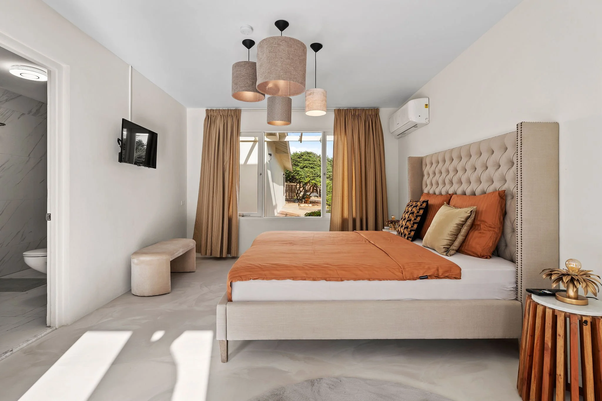 A bedroom with a large bed with orange bedding and multiple pillows, a tufted headboard, beige curtains, a small side table with a decorative lamp, a wall-mounted TV, and an opening leading outside with trees visible. There's also a small upholstered bench and a partial view of a bathroom with marble walls.