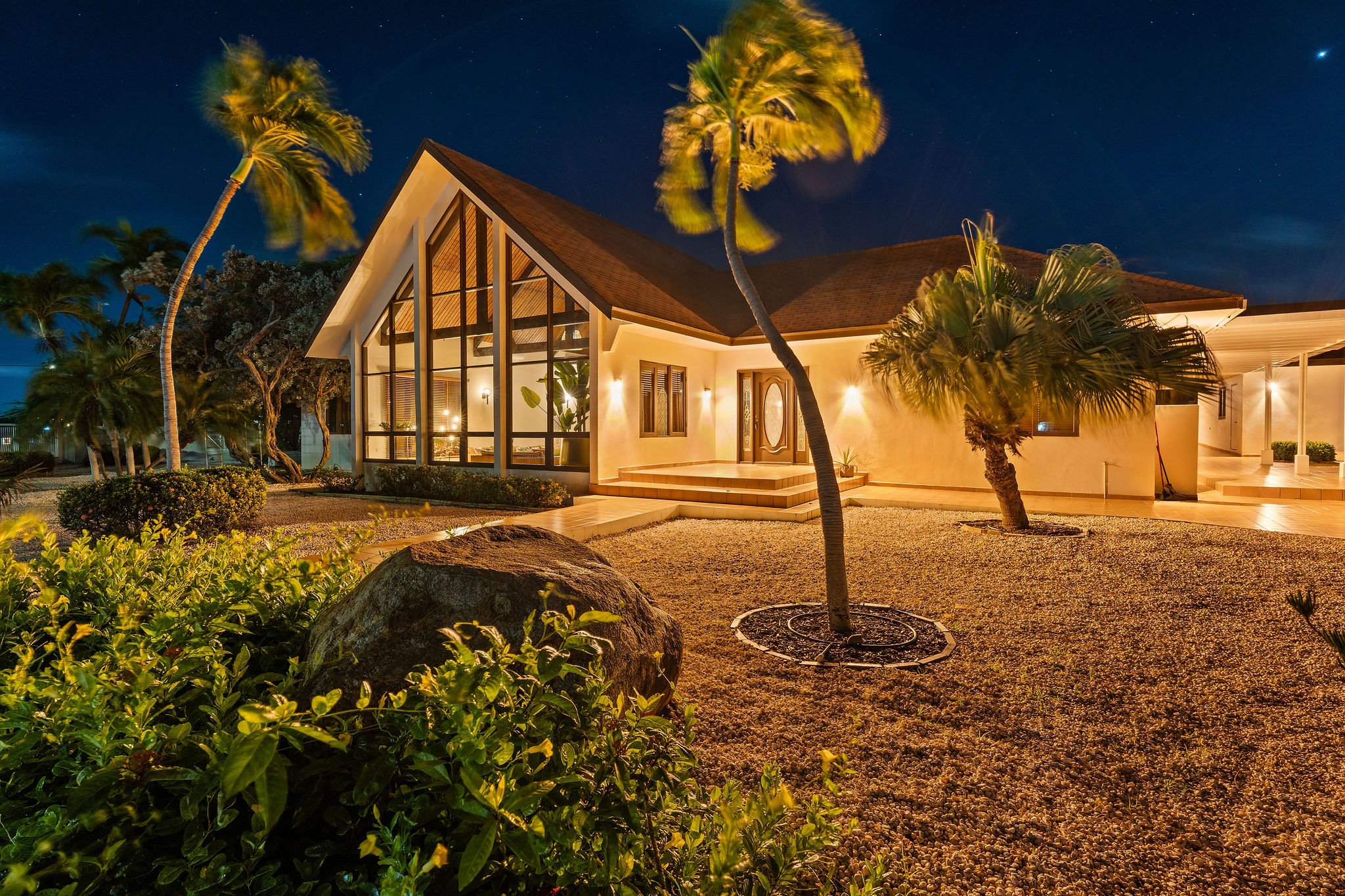 Night view of Villa casibari aruba with large glass windows and warm exterior lighting, surrounded by palm trees and landscaping, under a starry sky with visible stars.