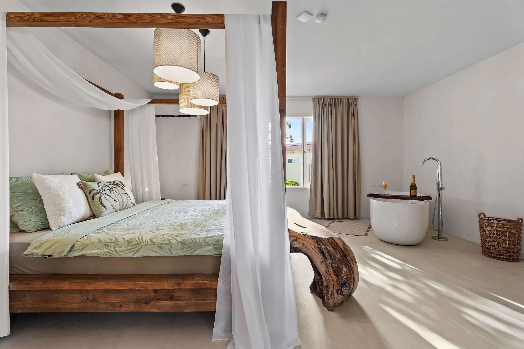 A cozy bedroom with a four-poster wooden bed, white bedding, and decorative pillows. Hanging lamps and curtains are visible, along with a window showing an outdoor view. The room includes a bathtub with a bottle of champagne and a glass, a wicker basket, and a rustic wooden side table with a natural wood log piece as a support.