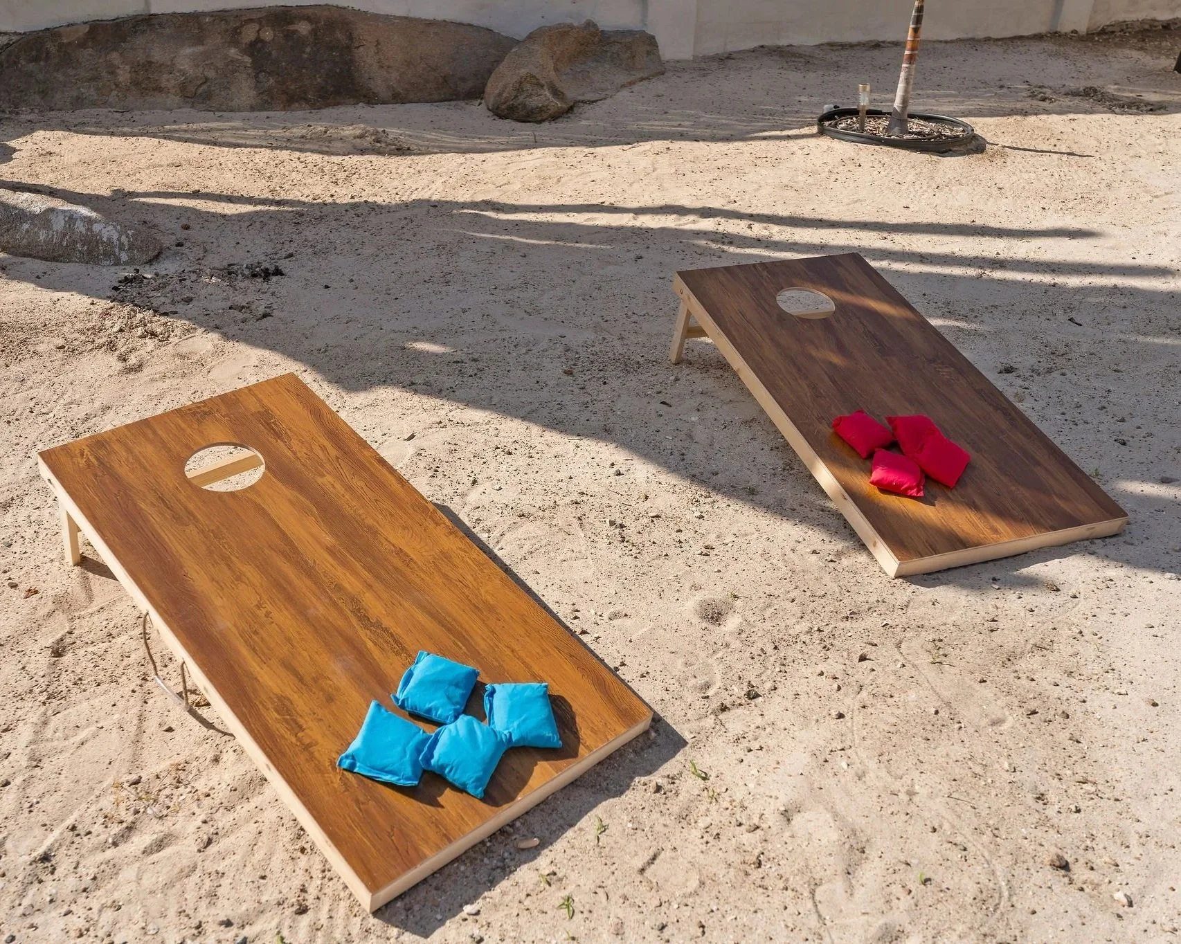 Two wooden cornhole boards with red and blue bean bags on a sandy outdoor surface, with shadows cast on the ground and rocks in the background.