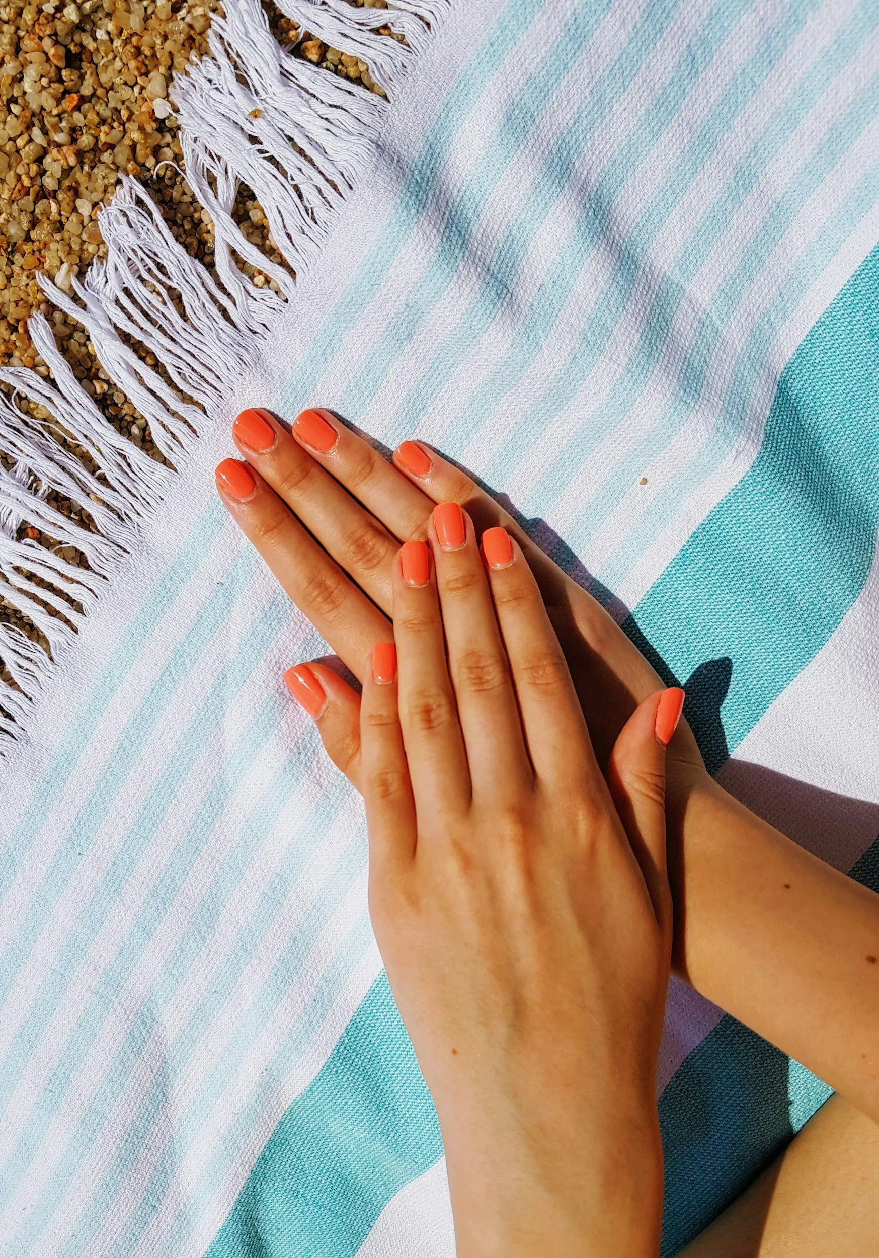 A person's hand with coral-colored nail polish resting on a striped blue and white towel on a sandy beach.