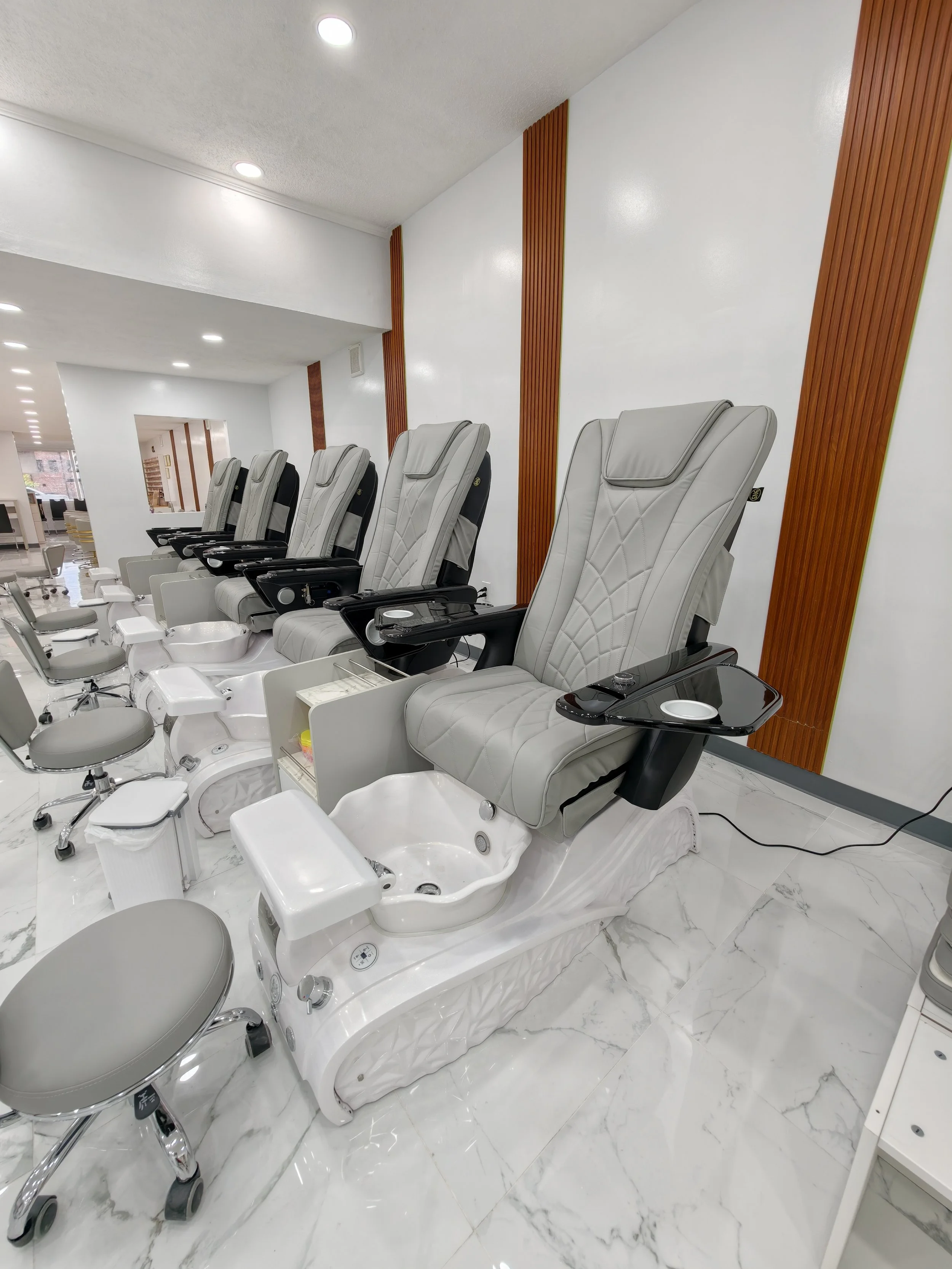 Multiple pedicure chairs in a salon, each with foot baths, arranged in a row along a white marble floor and white walls with wooden accents. Overhead lighting is visible.